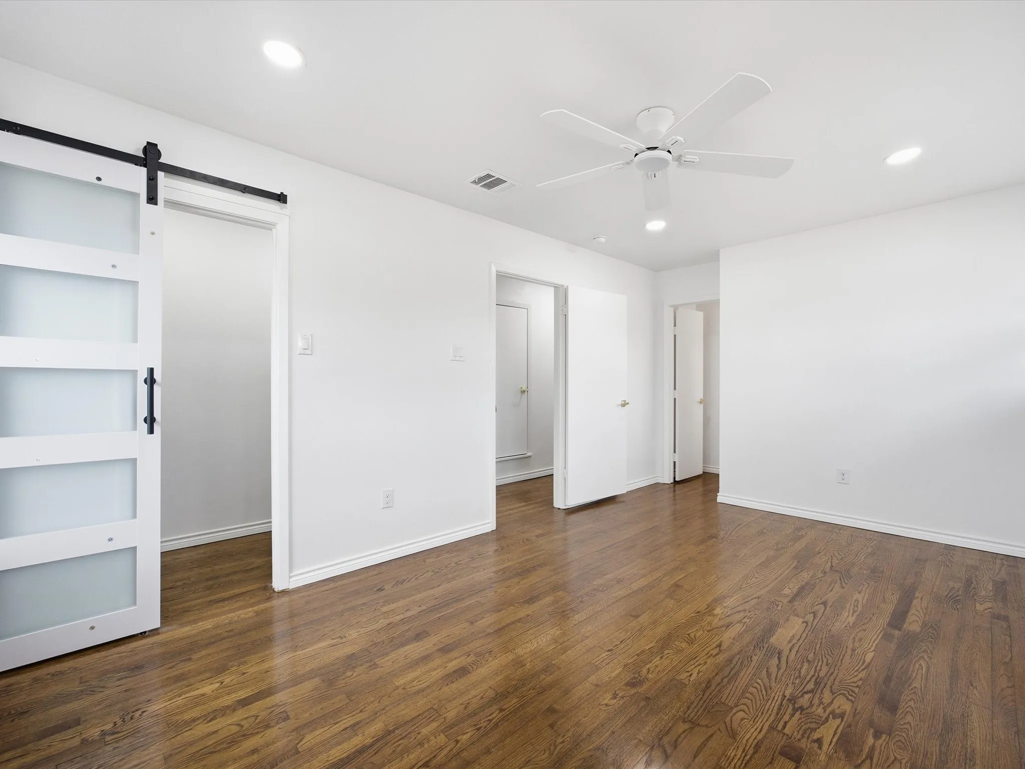 Wood-finish flooring, white walls, recessed lighting, ceiling fan, and barn door with frosted glass panels
