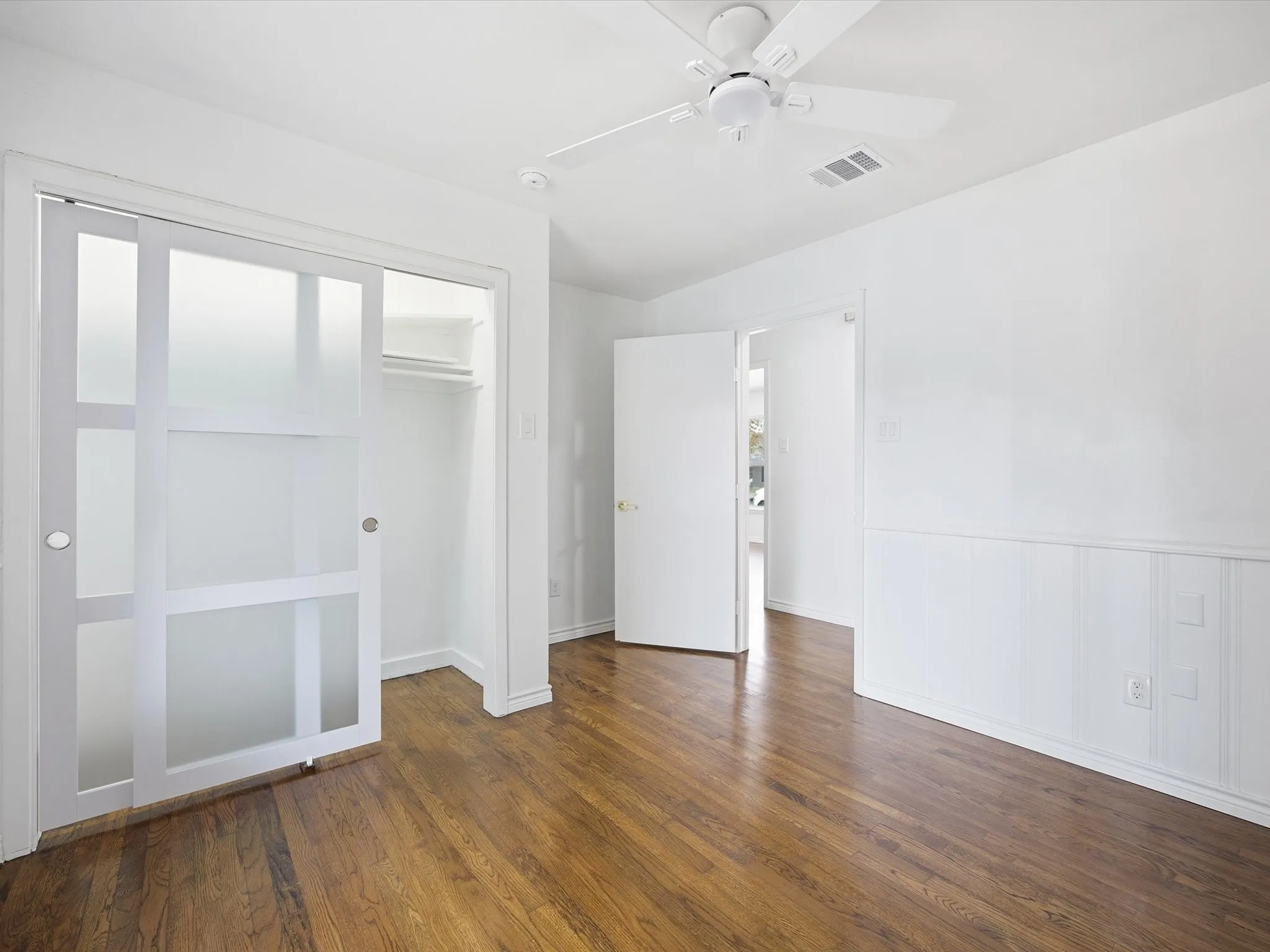 Wood-finish flooring throughout, white paneled wainscoting, white ceiling fan, and sliding frosted panel closet doors