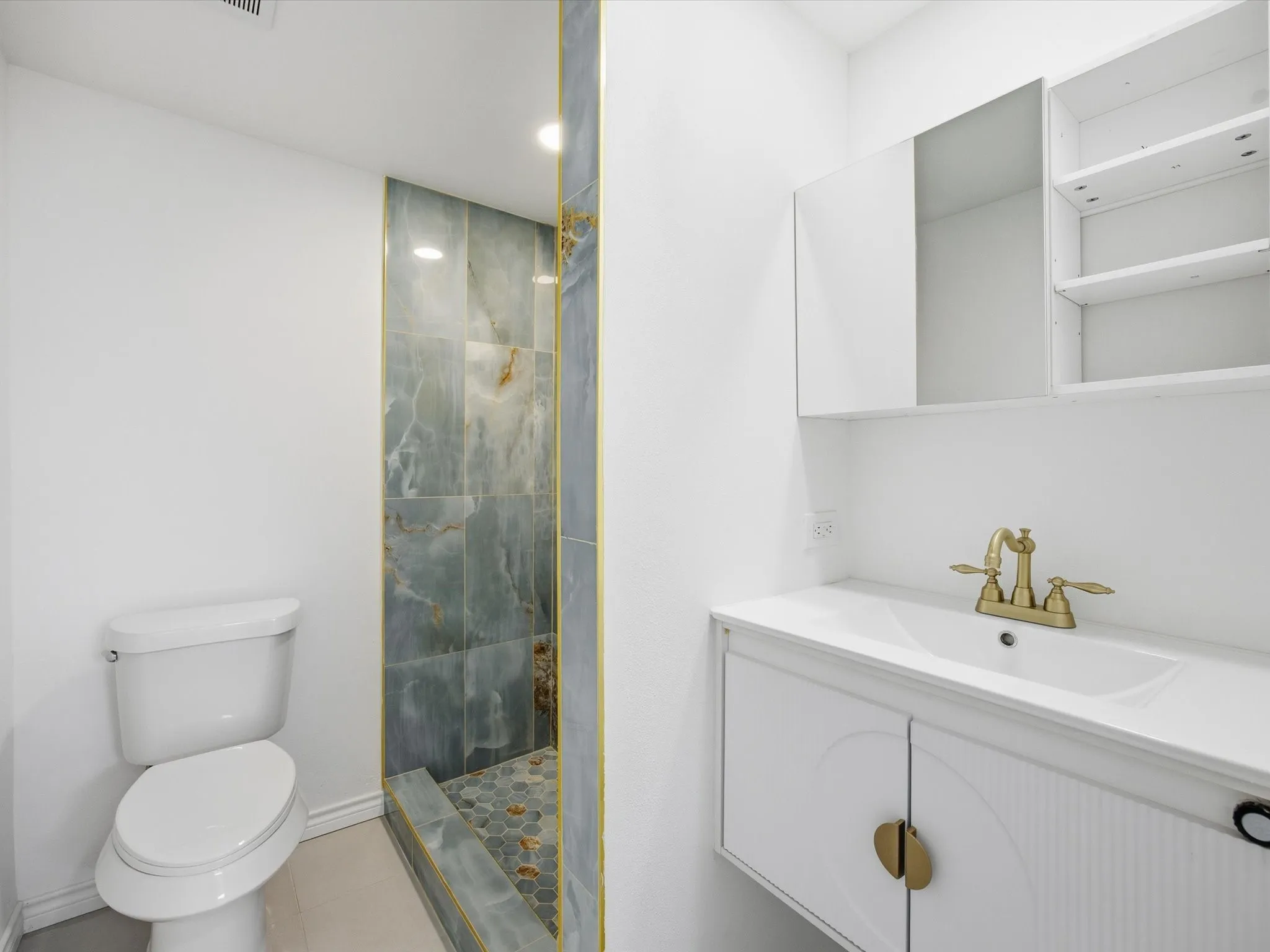 Vanity with integrated sink and brass-finish faucet, built-in medicine cabinet with open shelving, and a tiled shower with a mosaic floor