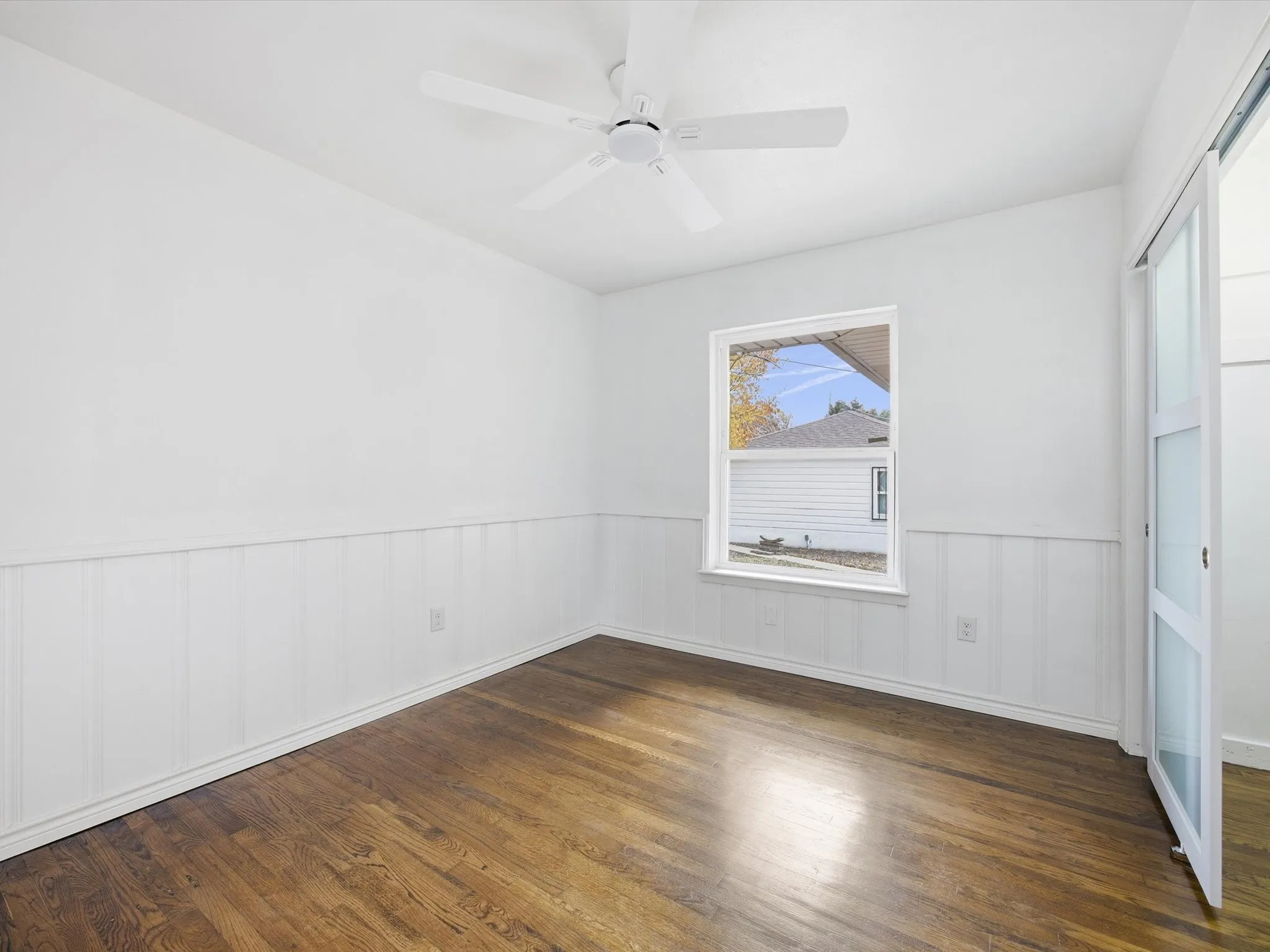Wood-finish flooring, white paneled wainscoting, white walls, and a white ceiling fan with integrated lighting