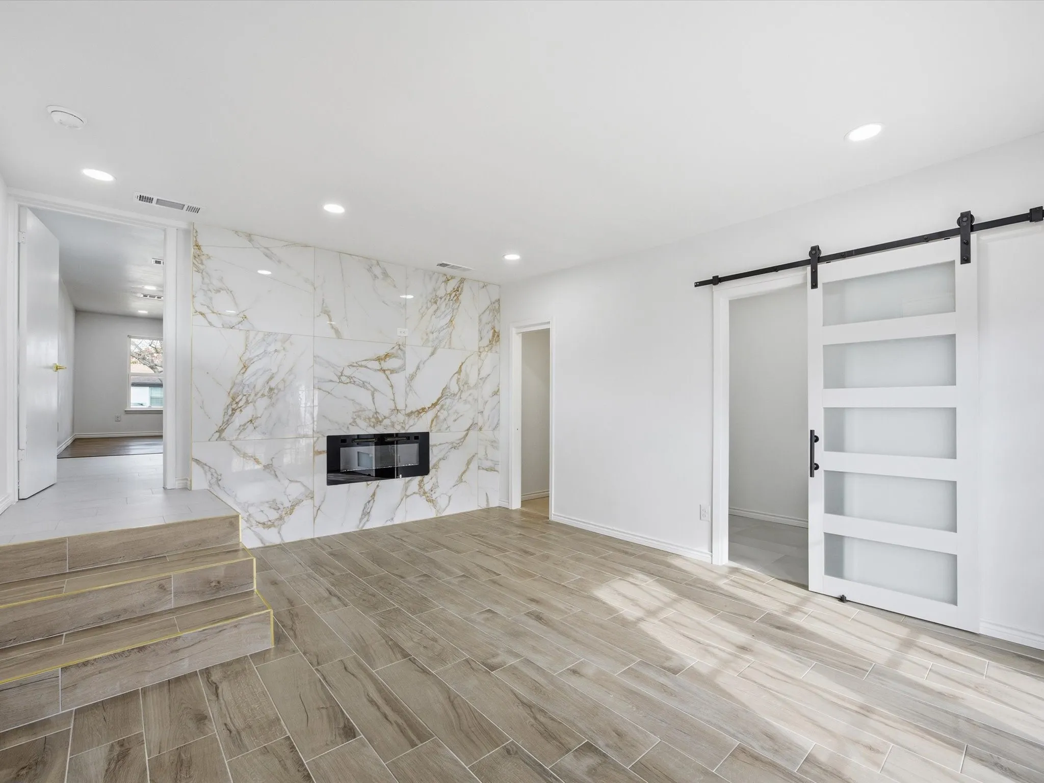 Porcelain tile flooring, recessed ceiling lighting, electric fireplace with a marble-look surround, and a white barn door with frosted panels