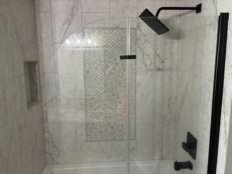 Bathtub shower featuring large-format tile, glass shower enclosure, matte black fixtures, and a decorative tile accent
