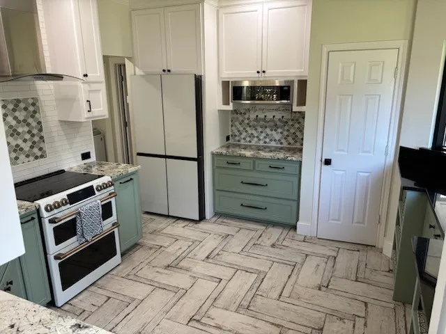 Herringbone pattern wood-finish flooring, white upper cabinetry, sage green lower cabinetry, and light-toned countertops with built in refrigerator