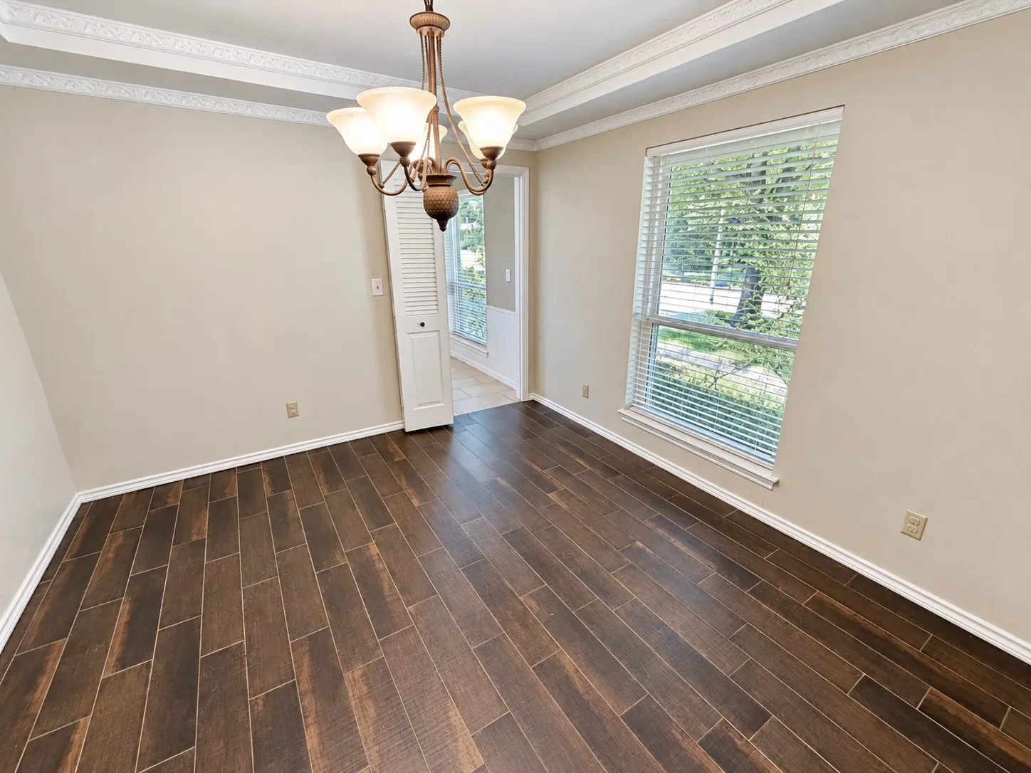 Wood-look tile flooring, overhead chandelier, decorative ceiling molding, and a large window with blinds