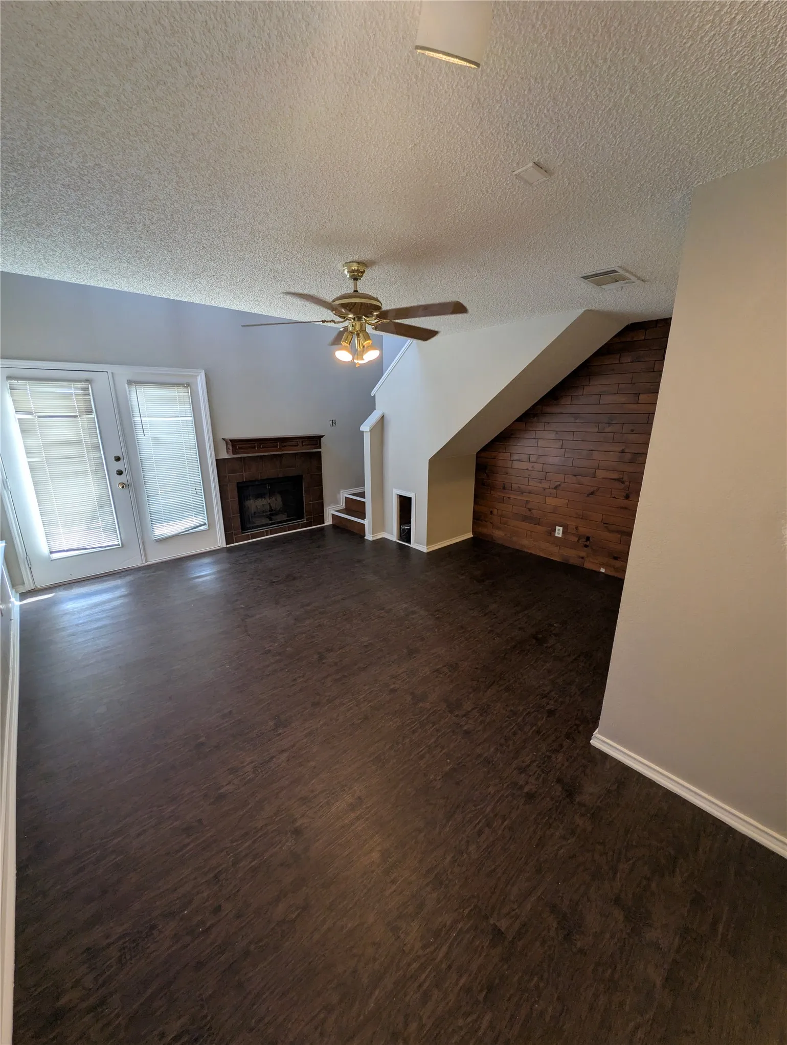 Dark wood-finish flooring throughout, a ceiling fan with integrated lighting, a fireplace with a wood mantle, and a wood-paneled accent wall