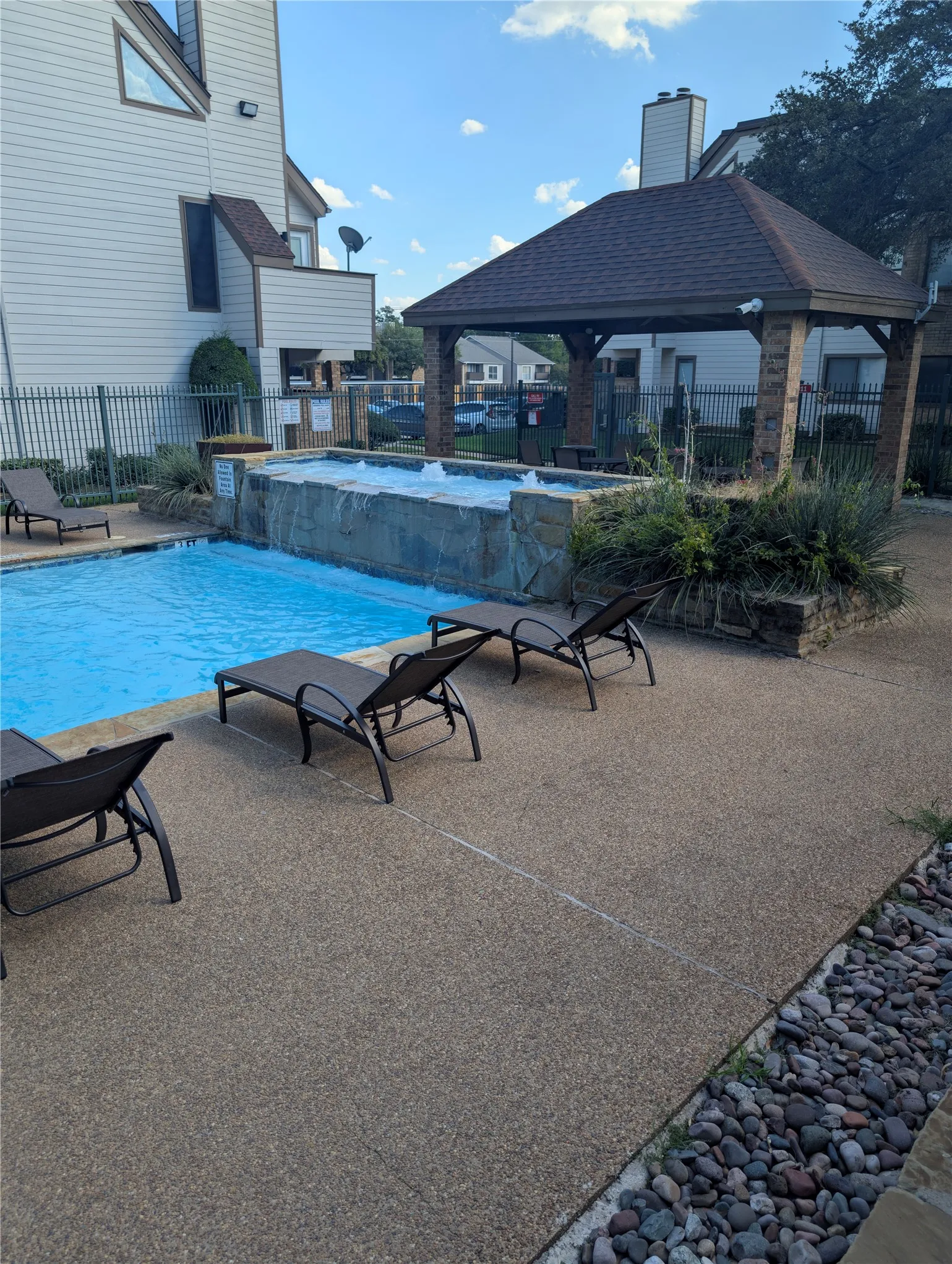 Resort-style pool area featuring a built-in spa with waterfall, stone-clad perimeter, and an adjacent brick-columned gazebo with a shingled roof
