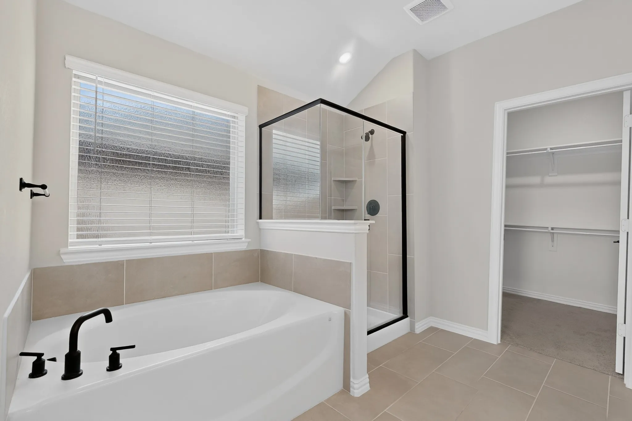 Soaking tub with matte black fixtures, separate enclosed shower with tiled walls and built-in shelving, tiled flooring, and a walk-in closet with built-in shelving
