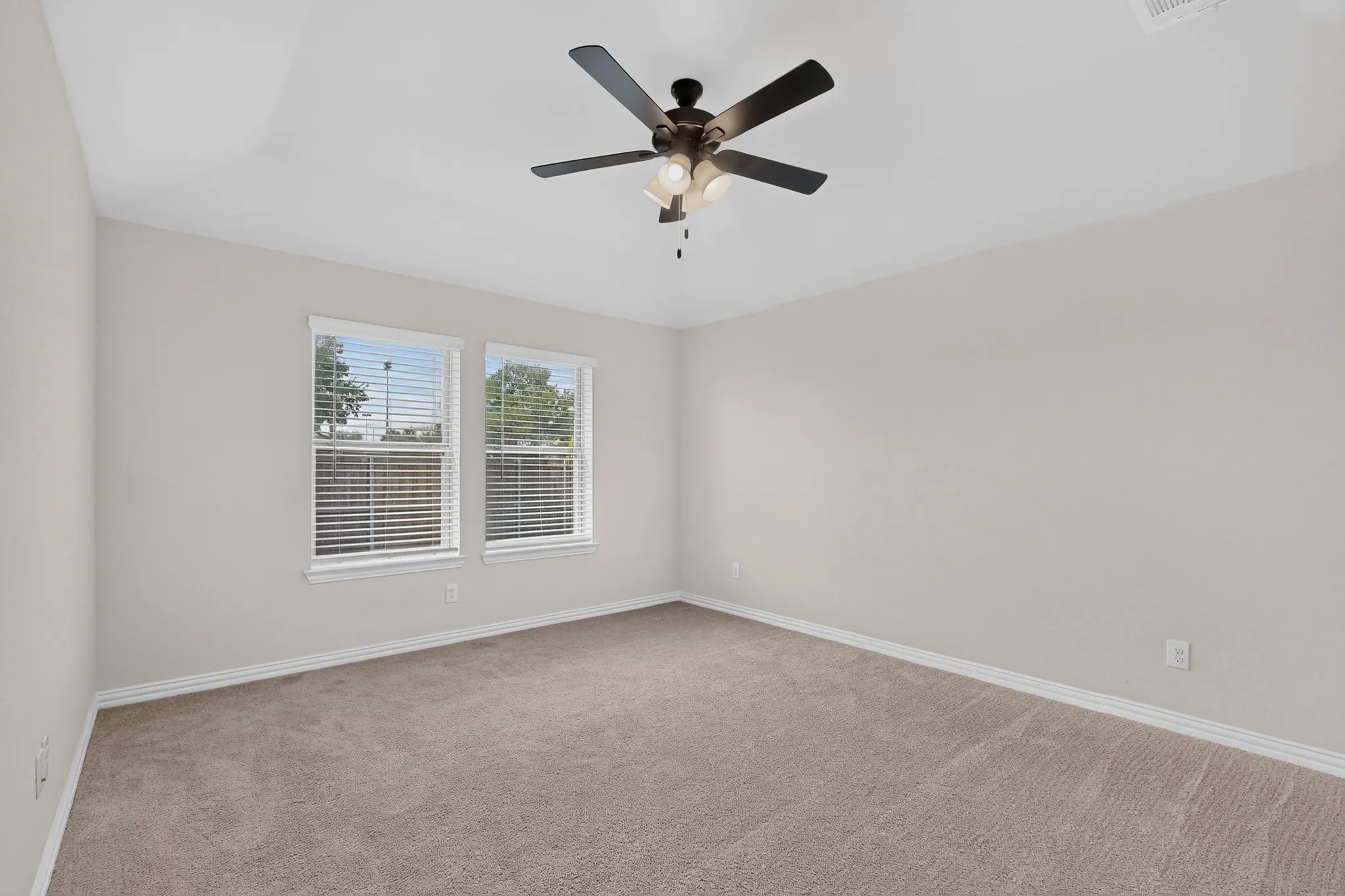Vaulted ceiling, two windows with blinds, ceiling fan with light fixture, neutral tone carpeting, and light-colored walls