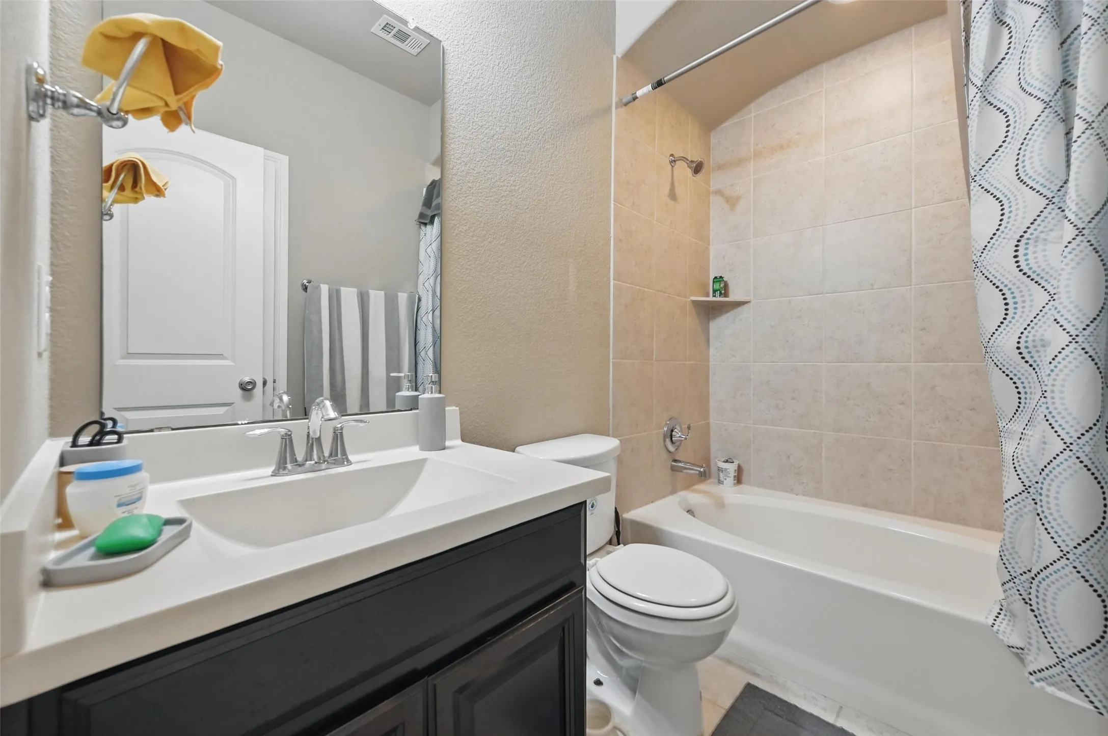 Vanity with an integrated sink and chrome faucet, dark wood-finish cabinetry, a full-width mirror, and a tiled shower-tub combination with a built-in shelf