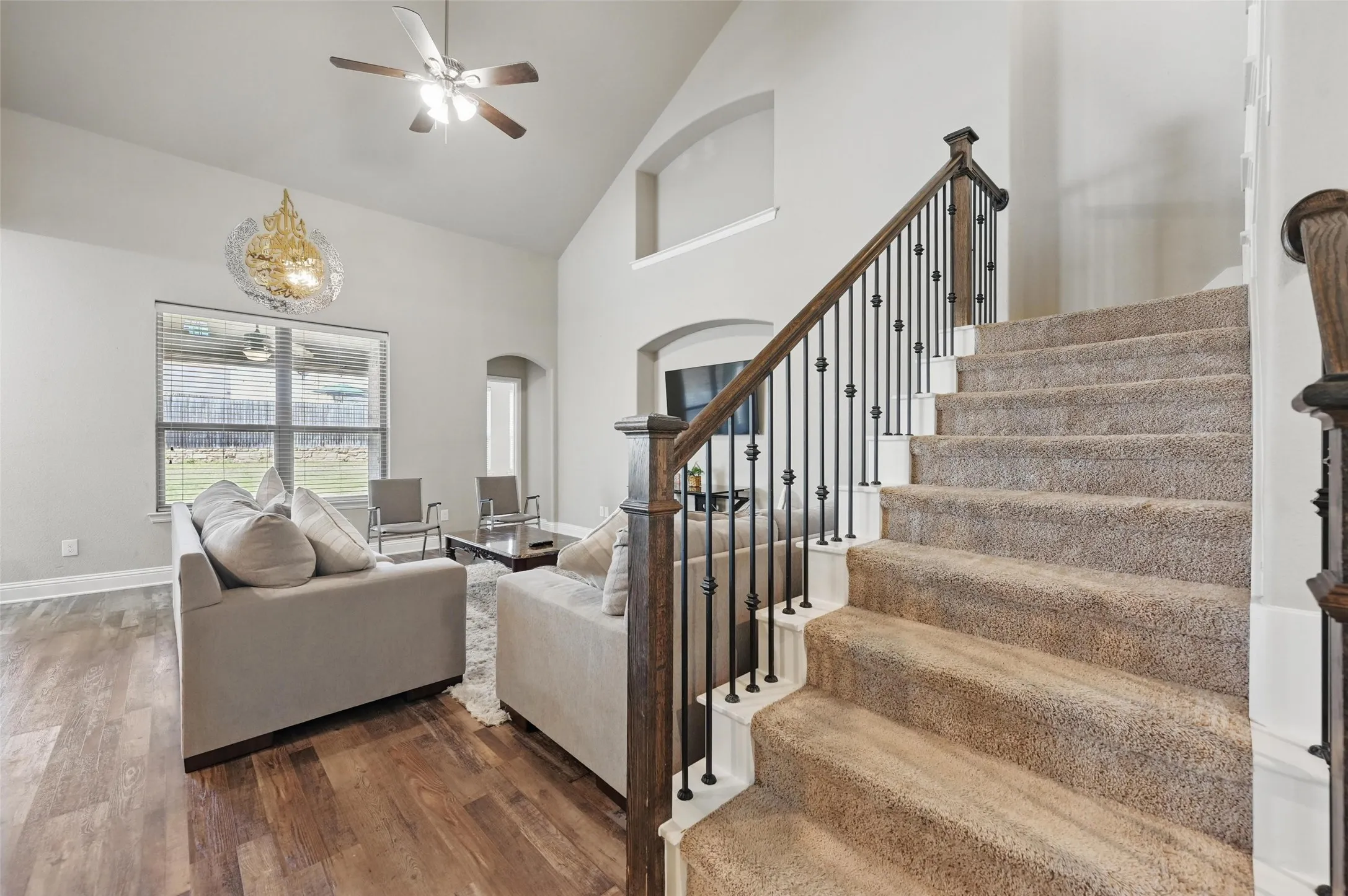 Vaulted ceiling with a ceiling fan, wood-finish flooring, and a carpeted staircase with wrought iron balusters