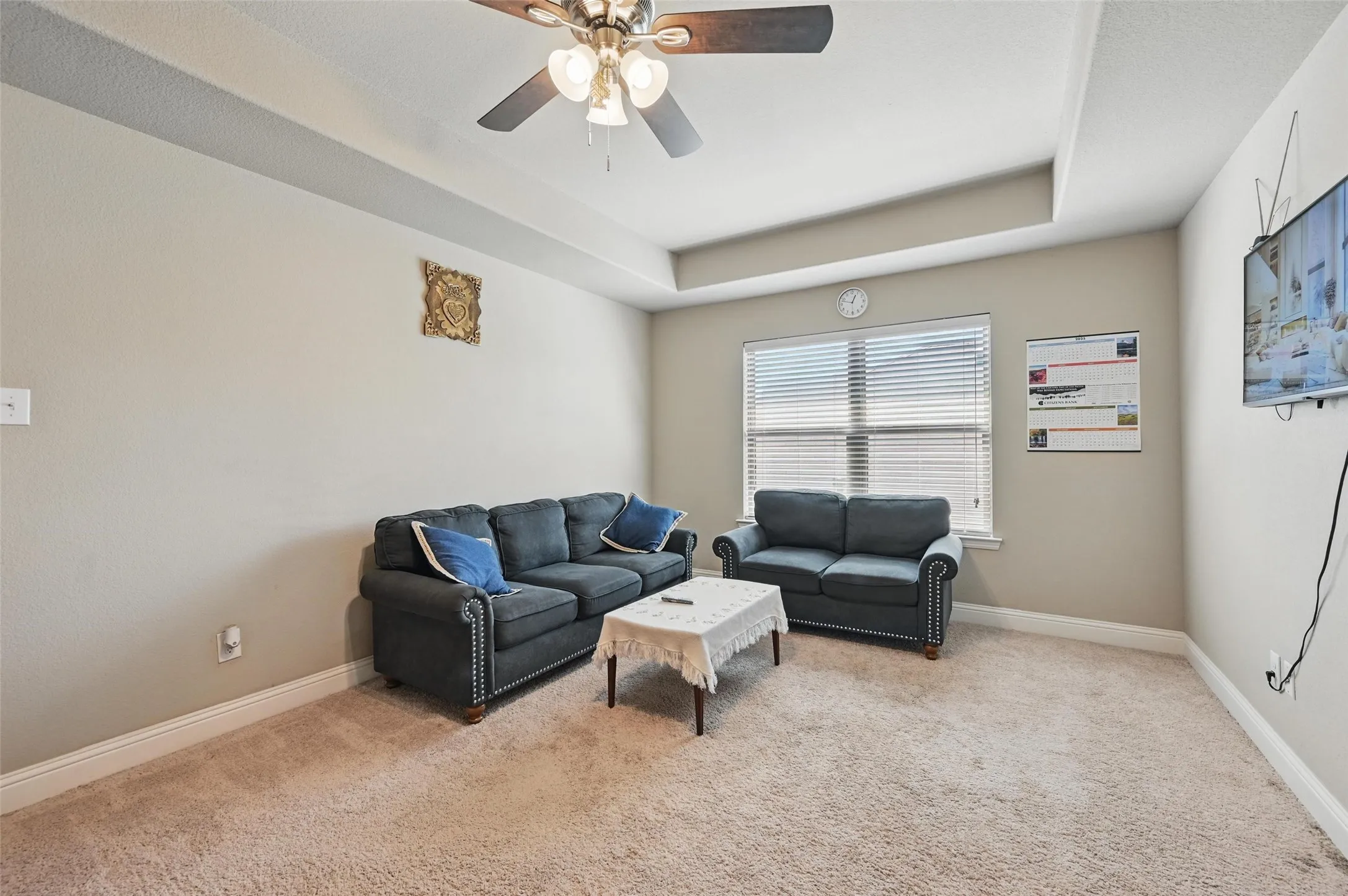 Carpeted interior space featuring a tray ceiling, ceiling fan with integrated lighting, and window with blinds