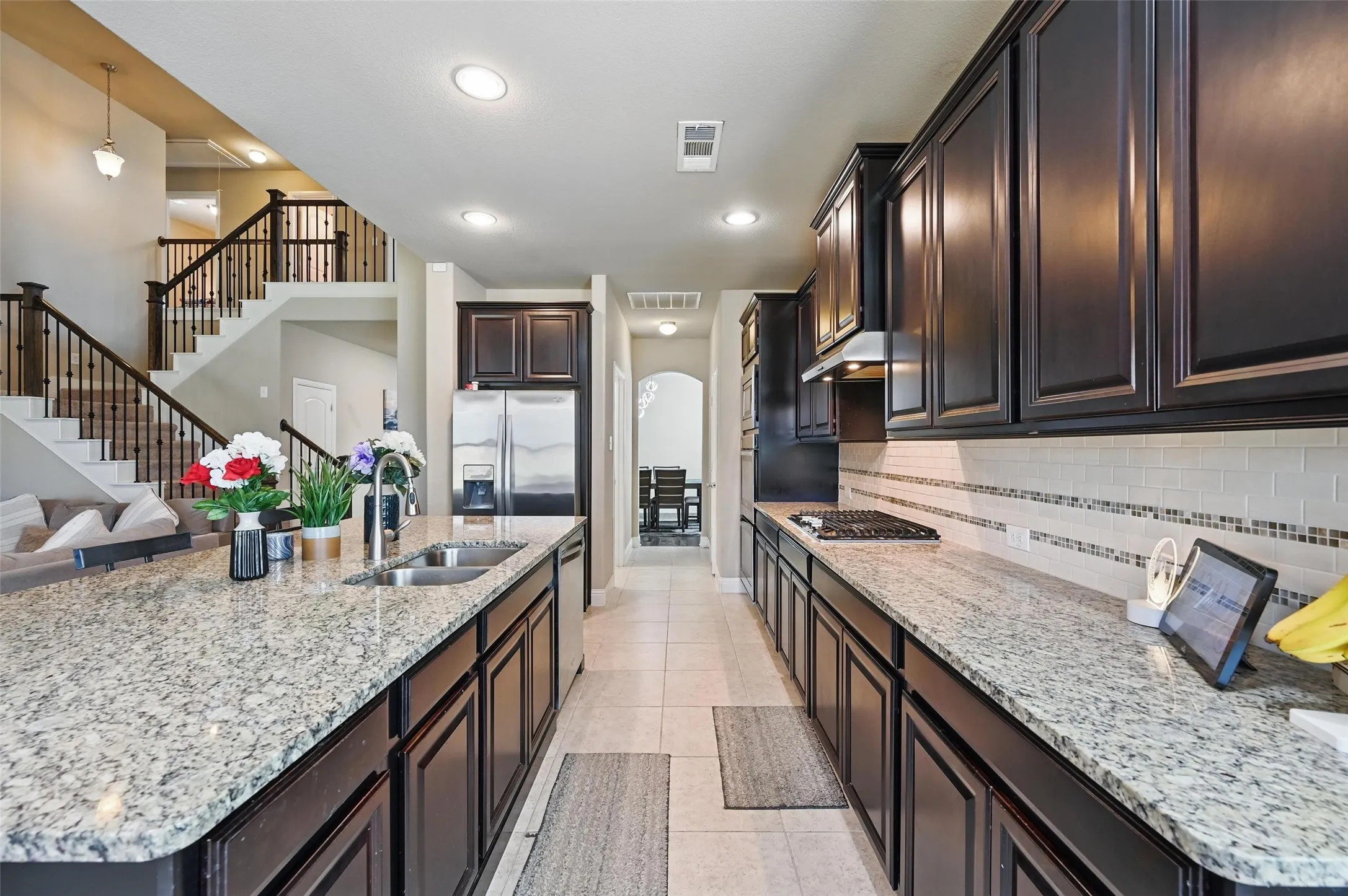 Kitchen featuring a large island with an undermount sink, dark wood cabinetry, light speckled countertops, stainless steel appliances, and a tile backsplash with mosaic accents