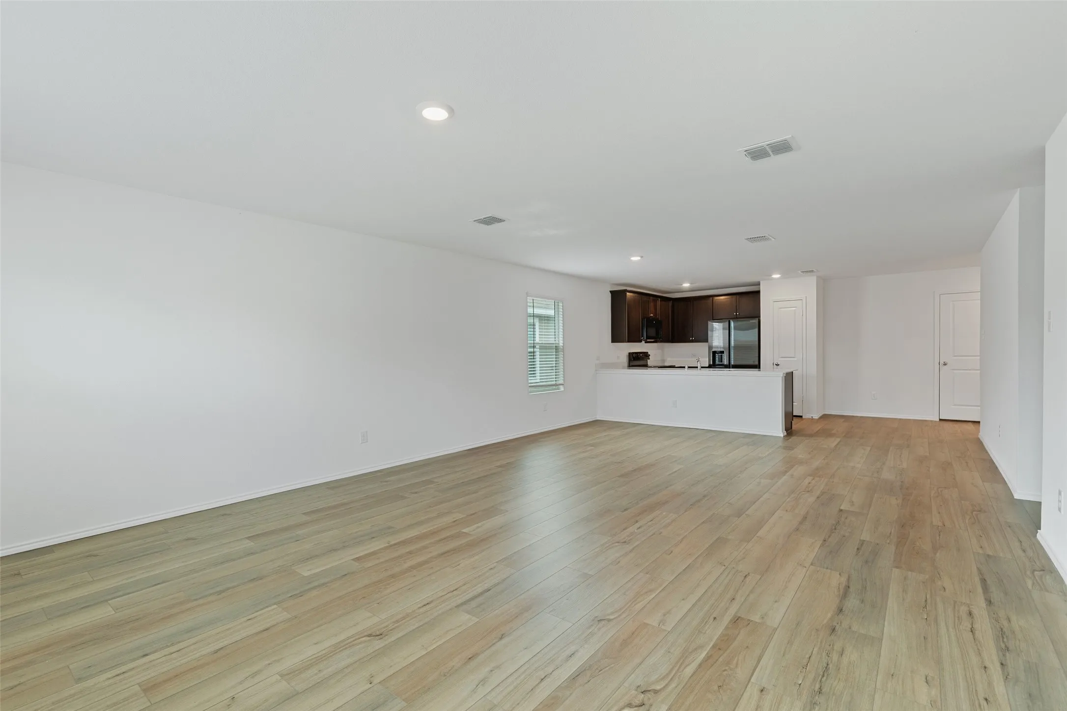 Spacious room with wood-finish flooring, recessed lighting, and a kitchen featuring dark wood cabinetry and a stainless steel refrigerator