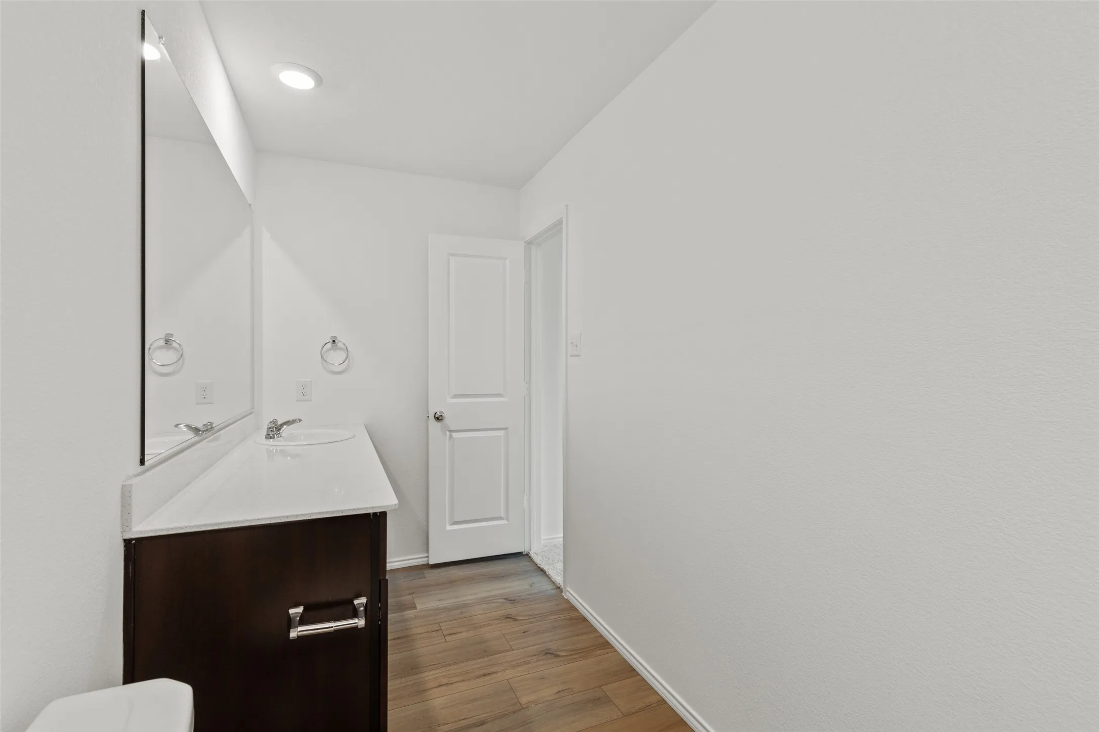 White walls, wood-finish flooring, dark wood-tone vanity, recessed lighting, and a white countertop with an integrated sink