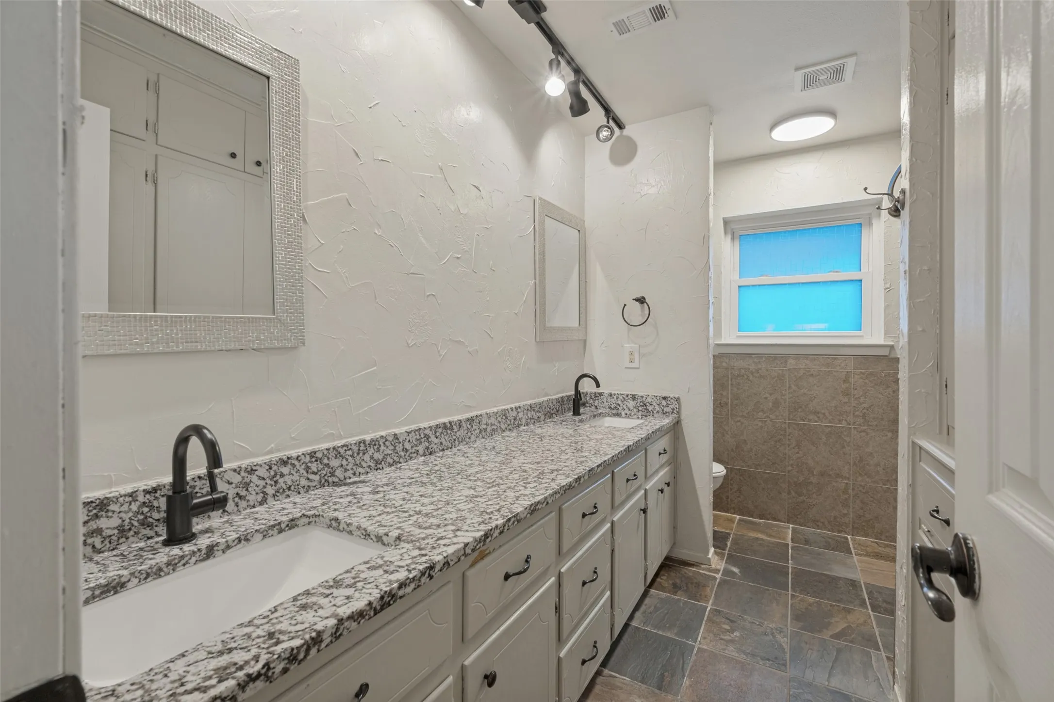 Dual sink vanity with granite countertop, matte black fixtures, built-in cabinetry, and tile flooring