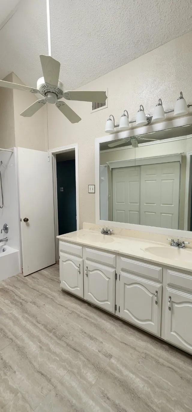 Vaulted ceiling with a ceiling fan, dual vanity with undermount sinks, wall-mounted light fixture, large vanity mirror, and wood-finish flooring