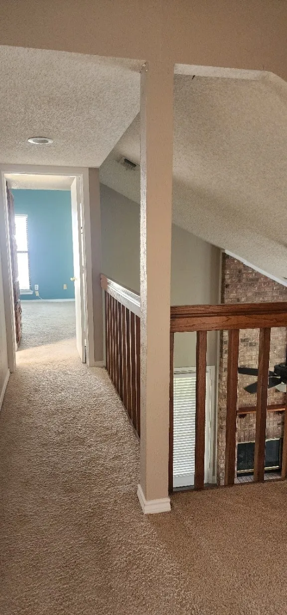 Carpeted hallway with a wood railing overlooking a lower space, featuring an exposed brick wall and a ceiling fan