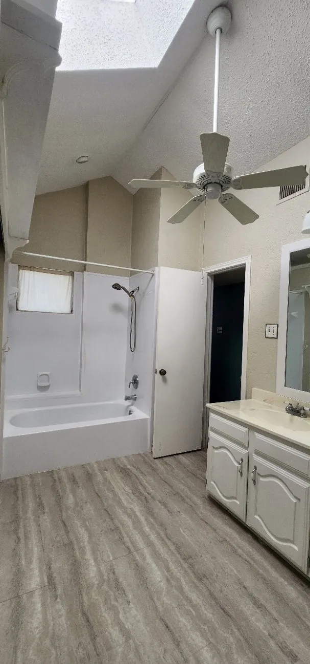Vaulted ceiling with skylight, ceiling fan, tub/shower combination, vanity with dual-door cabinetry, and wood-finish flooring