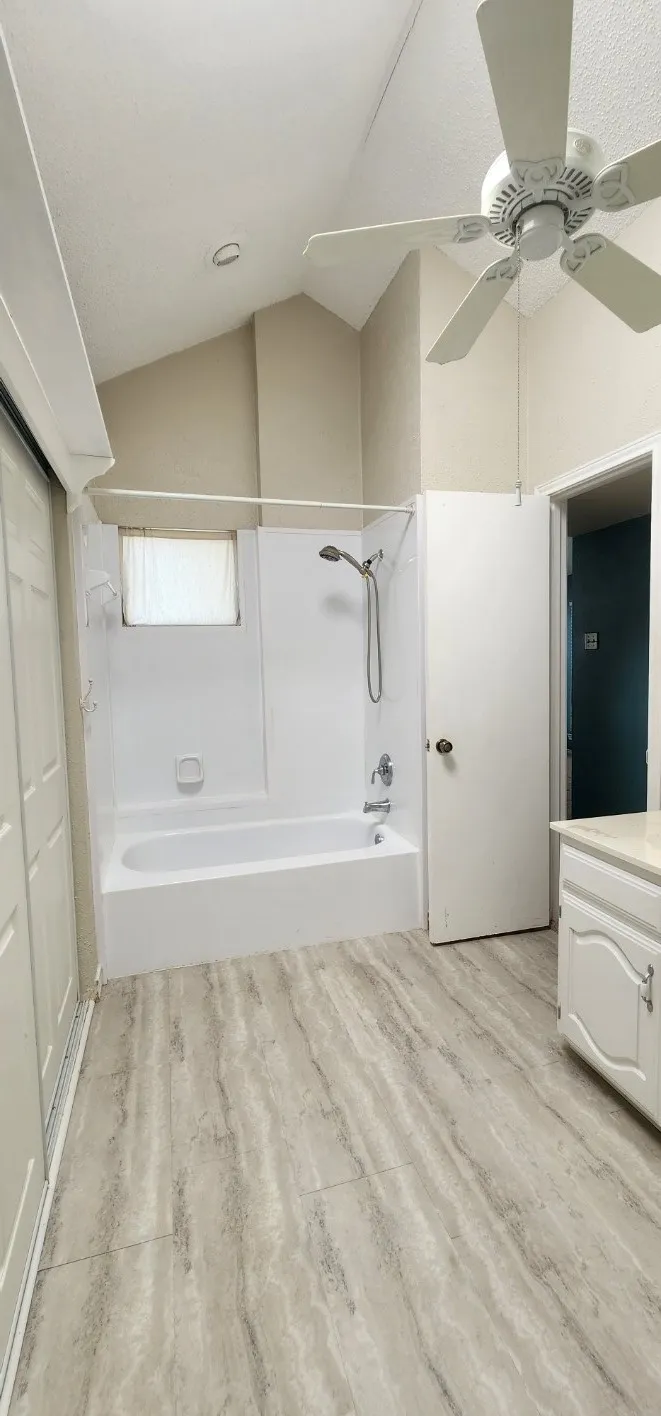 Vaulted ceiling with a ceiling fan, tub and shower combination, light-toned wood-finish flooring, white vanity with countertop, and sliding closet doors