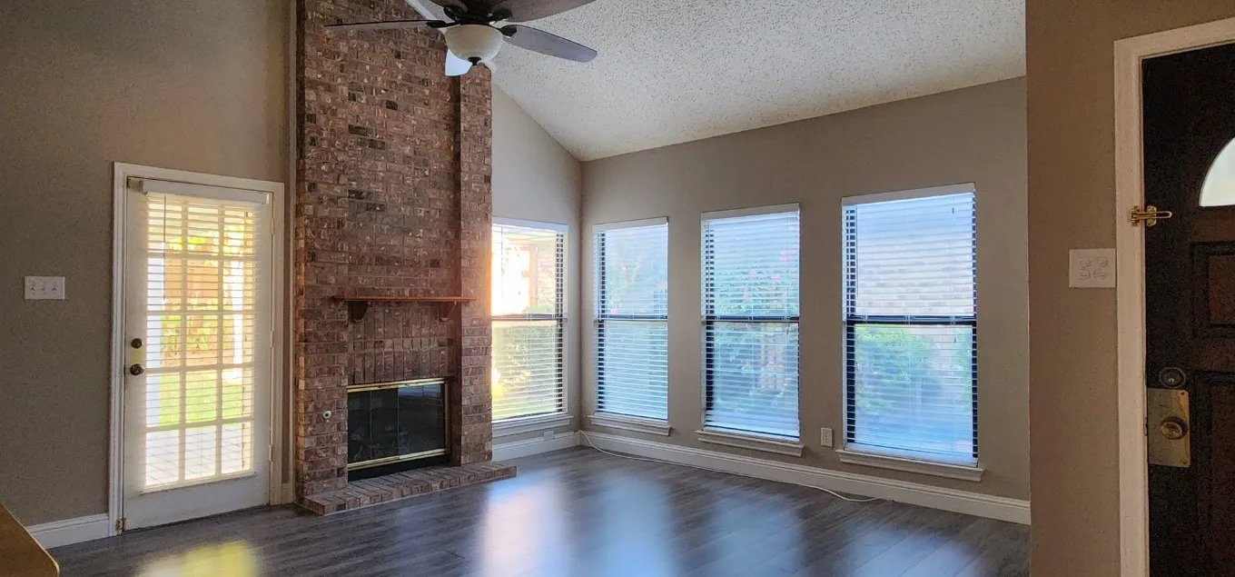 Vaulted ceiling, brick fireplace, wood-finish flooring, ceiling fan, and multiple windows