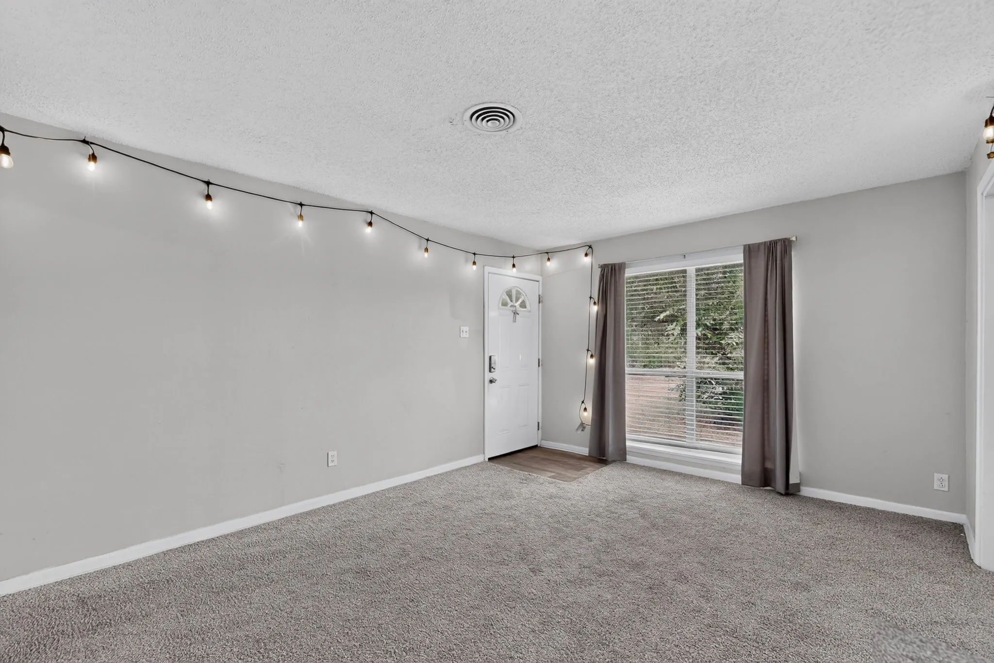 Neutral wall paint, textured ceiling, window with horizontal blinds, wood-finish flooring at entry, and soft carpet flooring