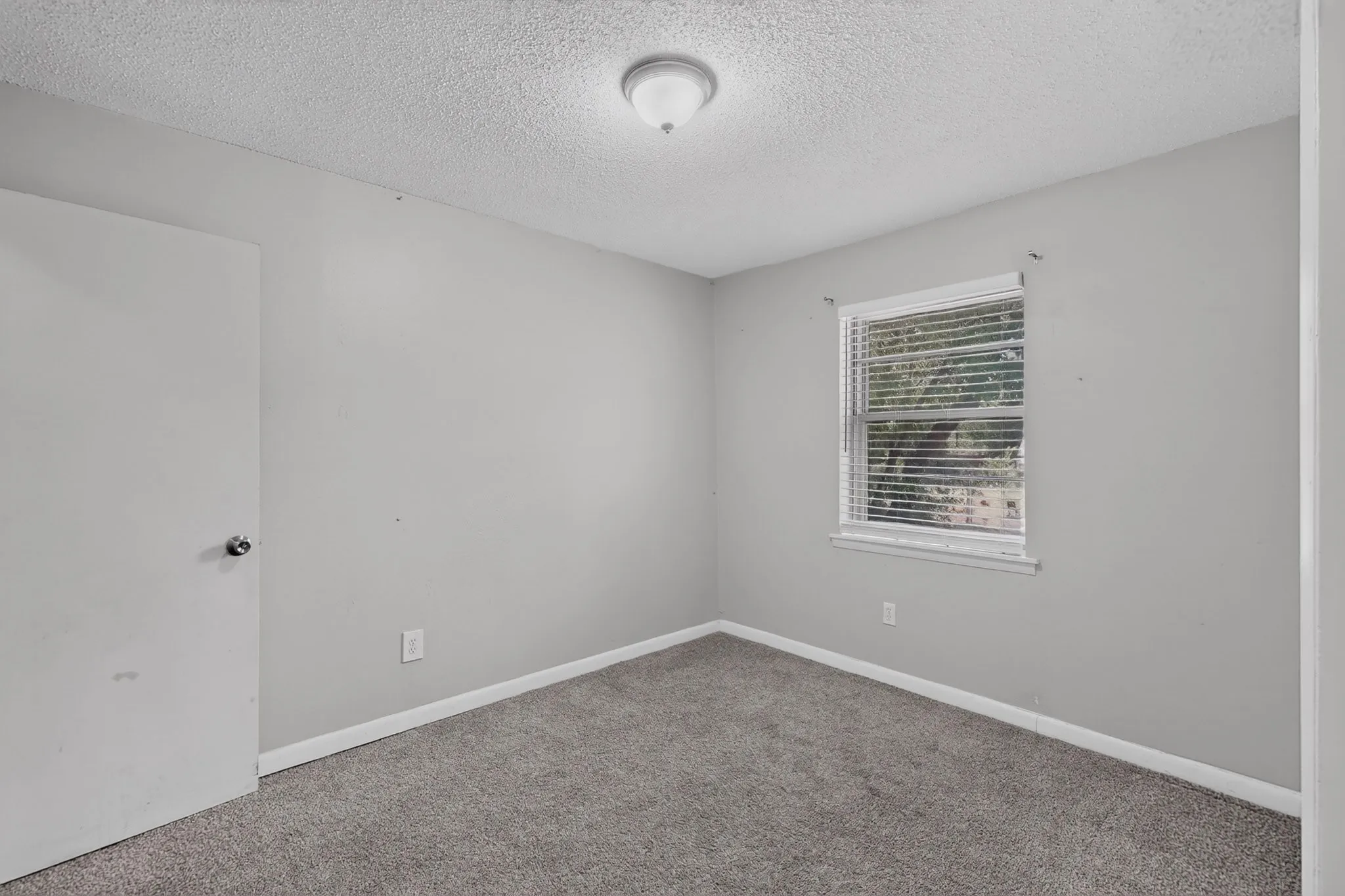 Neutral gray wall paint, textured ceiling, gray carpet flooring, window with blinds, flush mount ceiling light