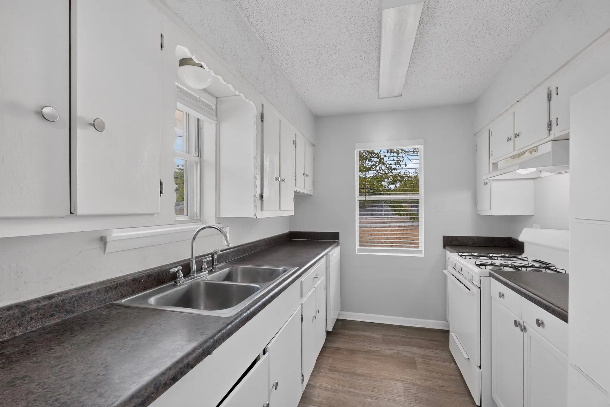 Galley kitchen featuring white cabinetry, dark countertops, a double basin stainless steel sink, and wood-finish flooring