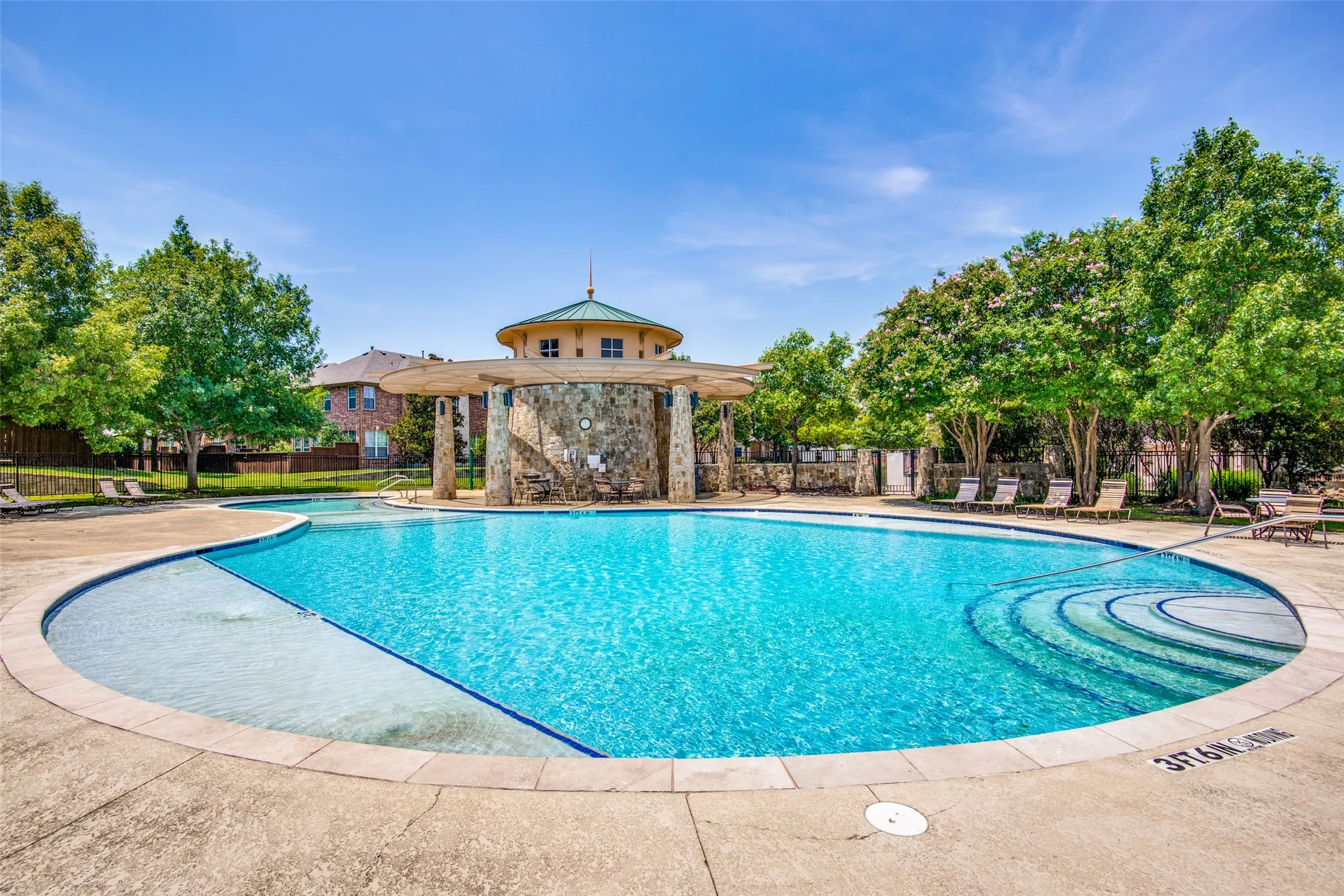 Resort-style swimming pool featuring a tanning ledge and integrated steps, surrounded by a concrete deck and mature trees