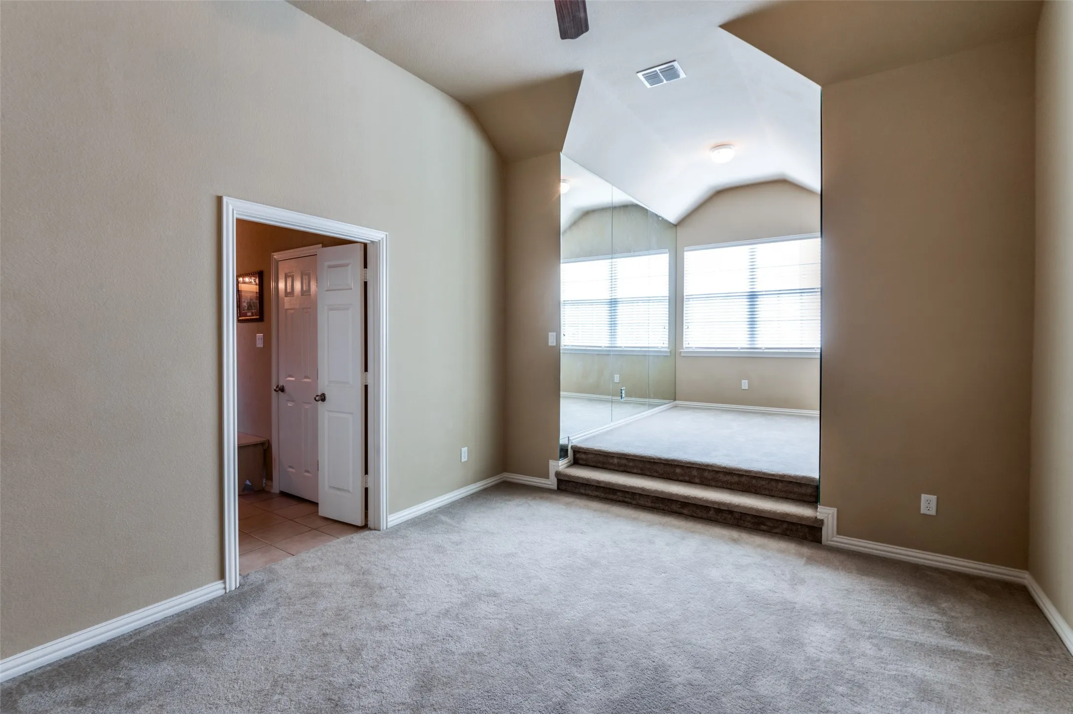 Carpeted room with vaulted ceilings and a two-step transition to an elevated space featuring three windows with blinds, a mirrored wall, and a ceiling light fixture