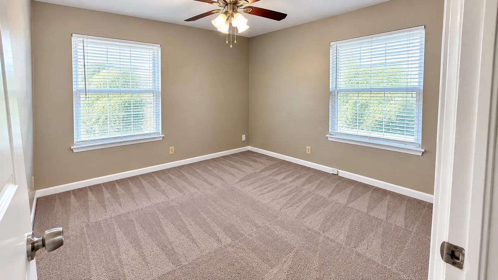 Neutral tone wall paint, dual windows with blinds, patterned carpet flooring, and a ceiling fan with integrated lighting