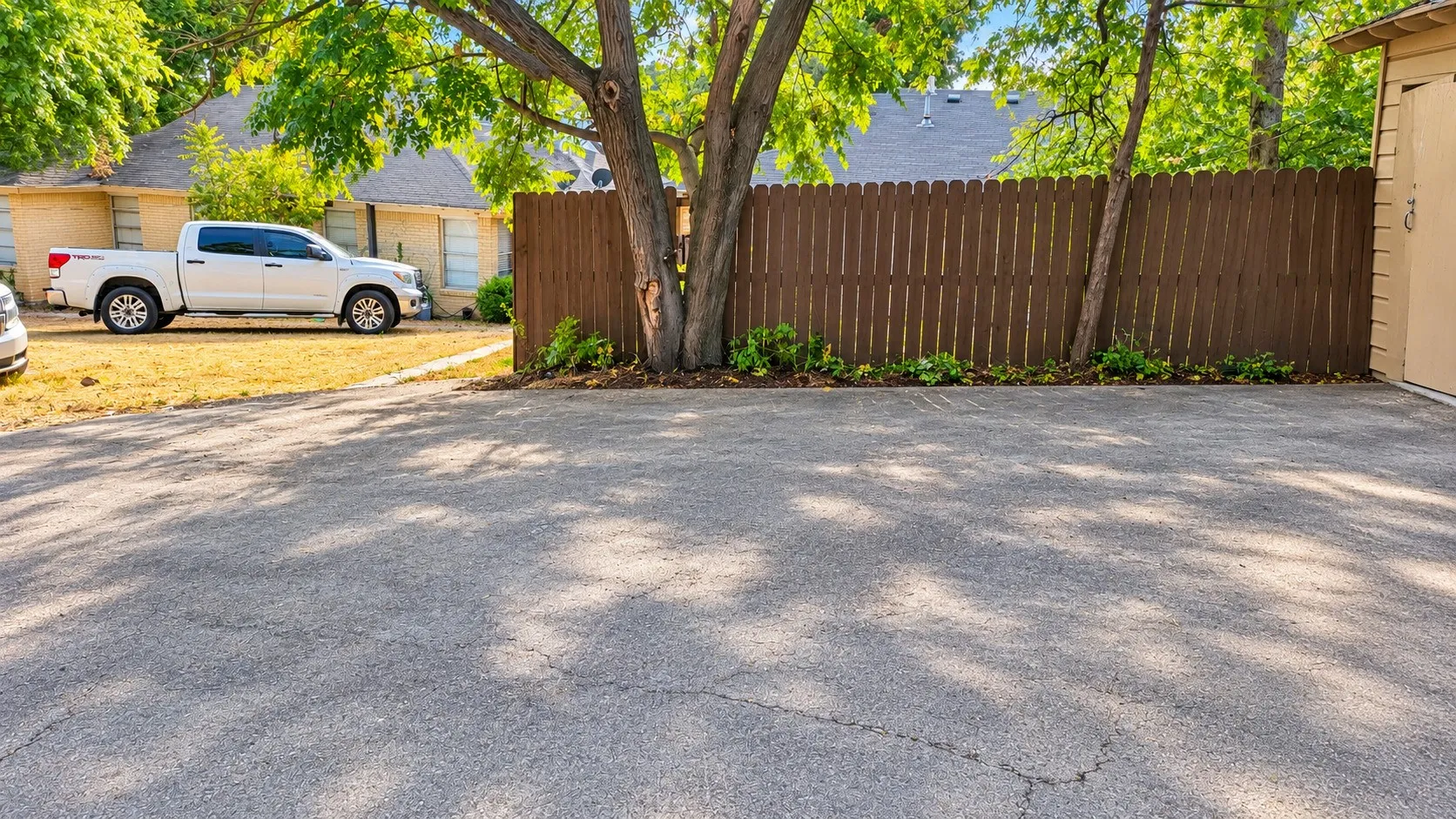 Paved driveway with mature trees, established landscaping, and a privacy fence