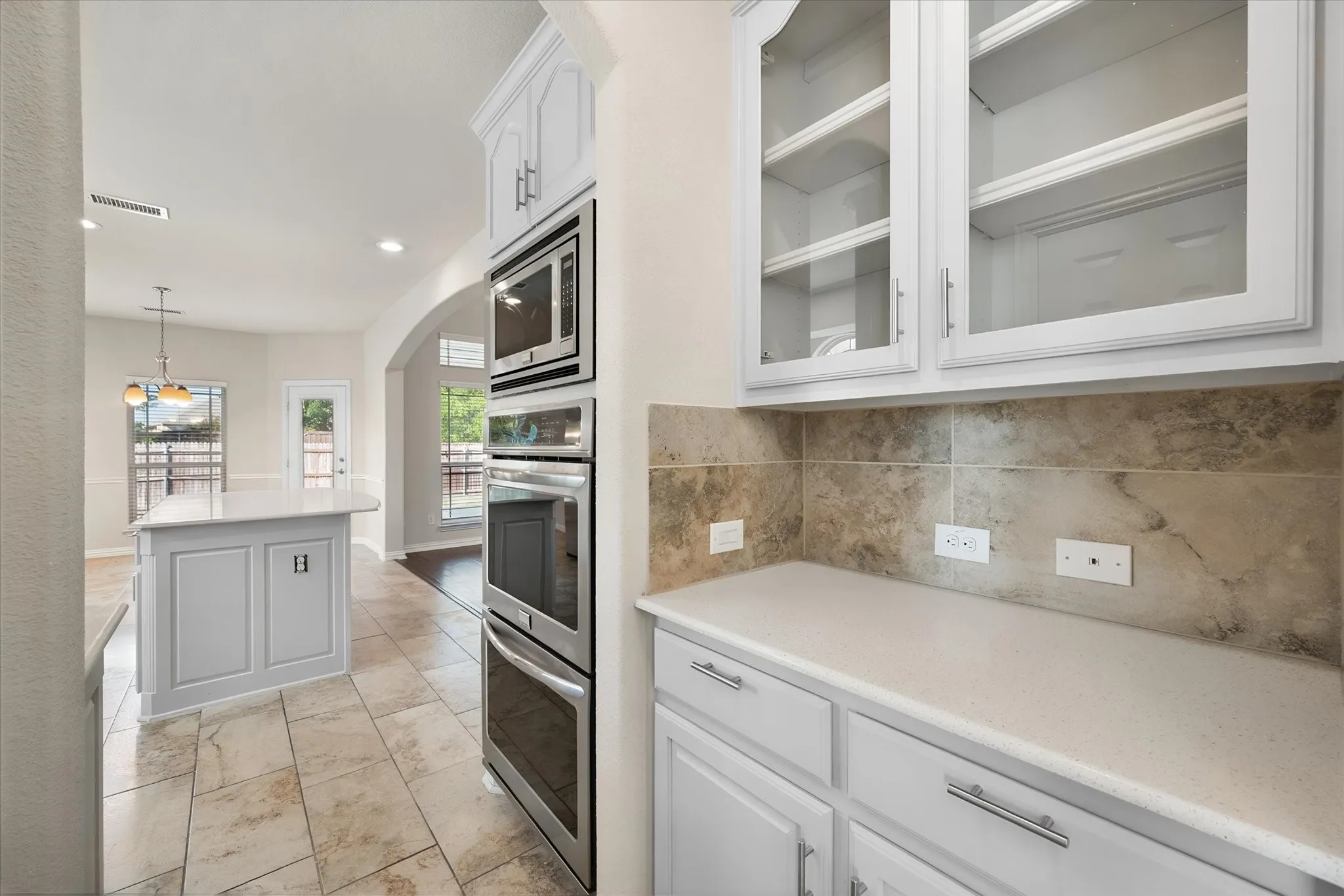 White cabinetry with glass-front uppers, light-toned countertop, stainless steel double oven, and tile backsplash
