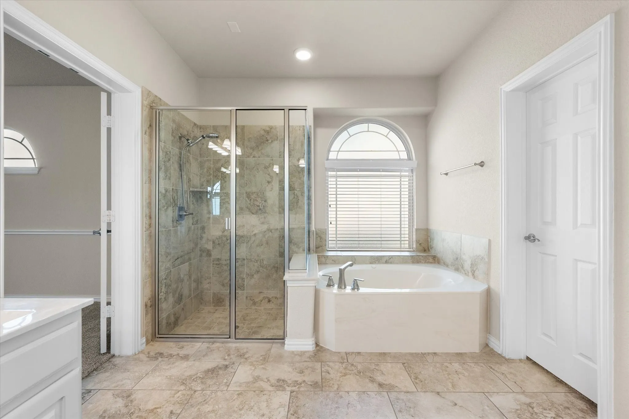 Tiled flooring throughout, corner soaking tub with deck-mounted fixtures, separate glass-enclosed shower with tiled walls, and an arched transom window with blinds