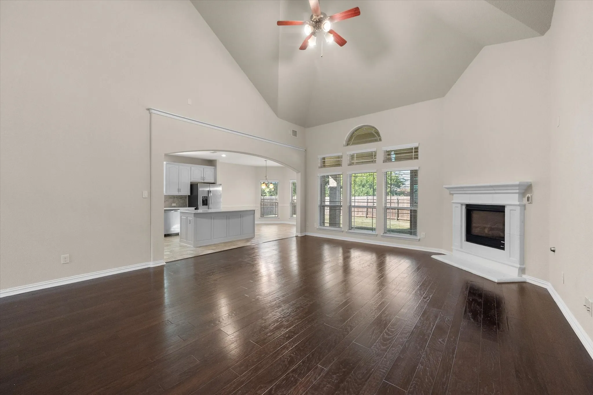 Vaulted ceiling with a ceiling fan, dark wood-finish flooring, white fireplace with hearth, arched entryway, and multiple windows