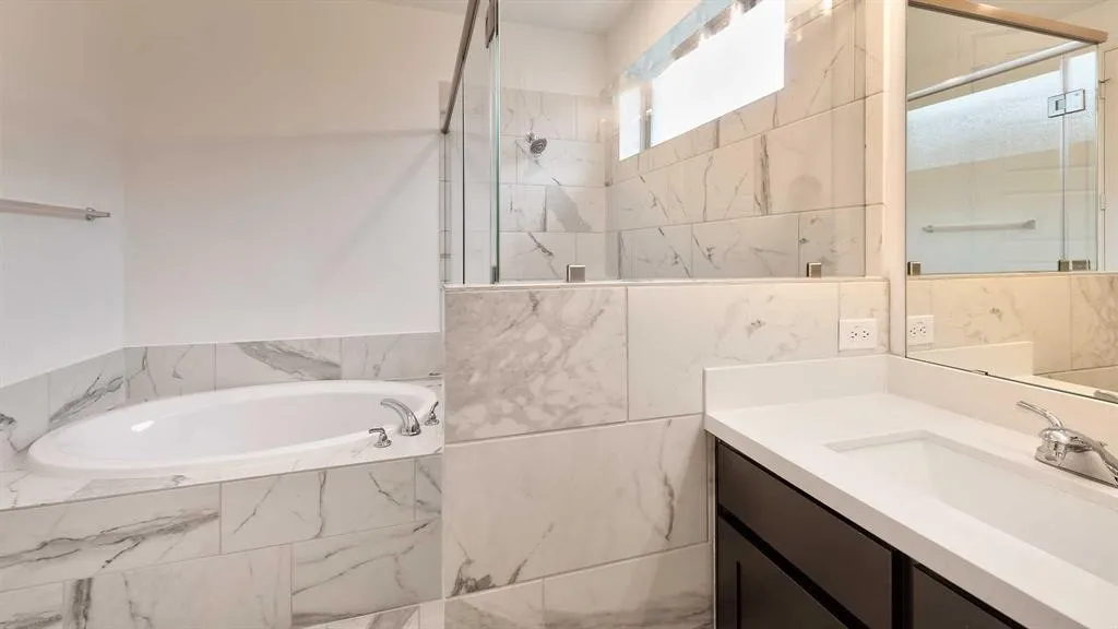 Soaking tub with polished chrome fixtures, walk-in shower with clear glass enclosure, extensive marble-look tile, single vanity with integrated sink, and dark wood-finish cabinetry