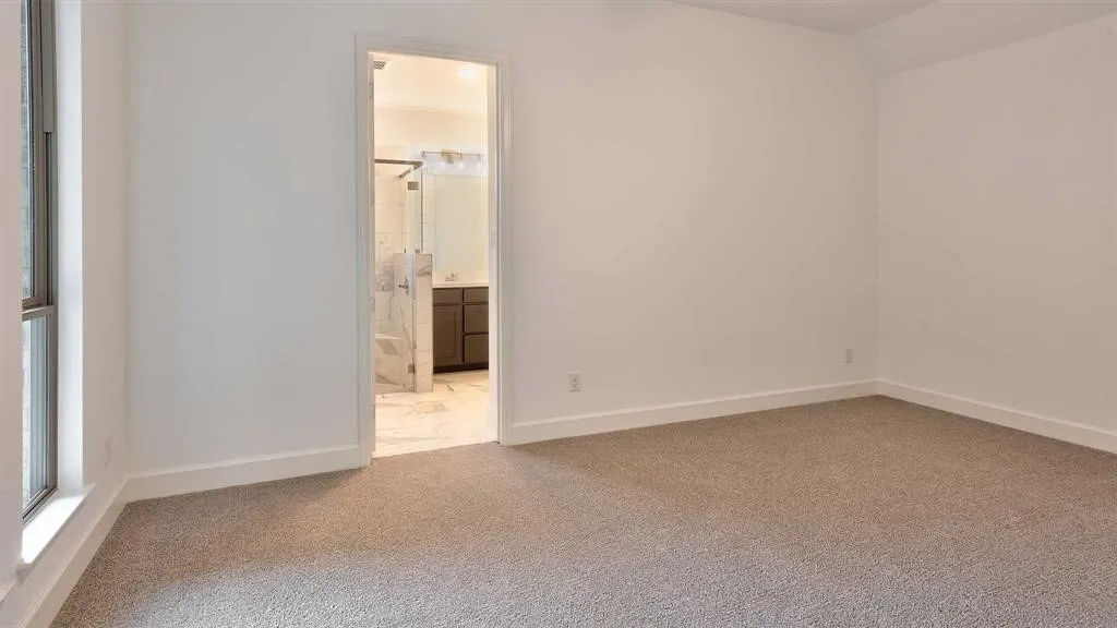 Carpeted bedroom with a window and a doorway leading to a bathroom featuring a glass-enclosed shower and a vanity with a dark-finished cabinet
