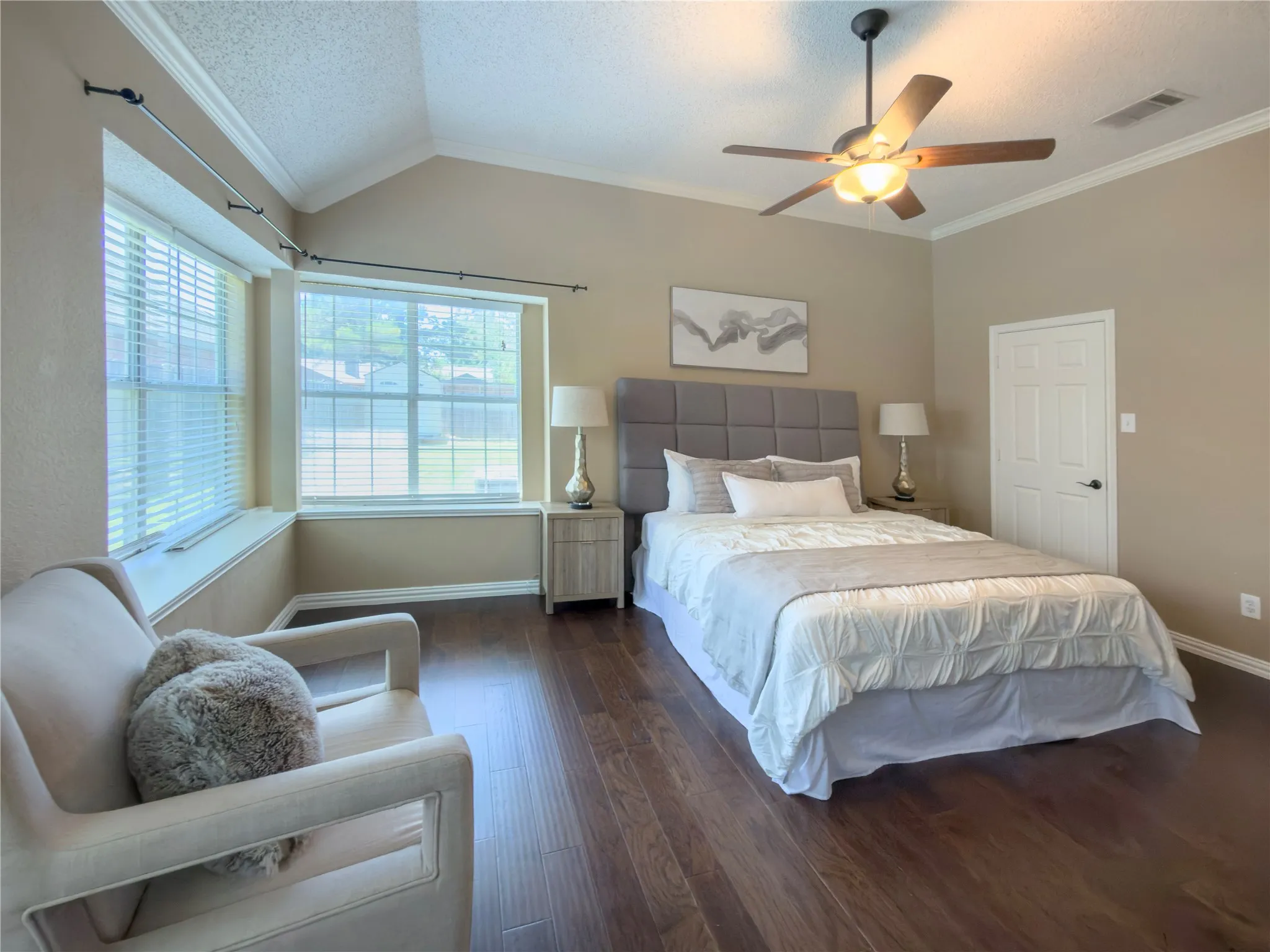 Vaulted ceiling with crown molding, wood-finish flooring, two large windows, ceiling fan with light fixture, and a paneled door
