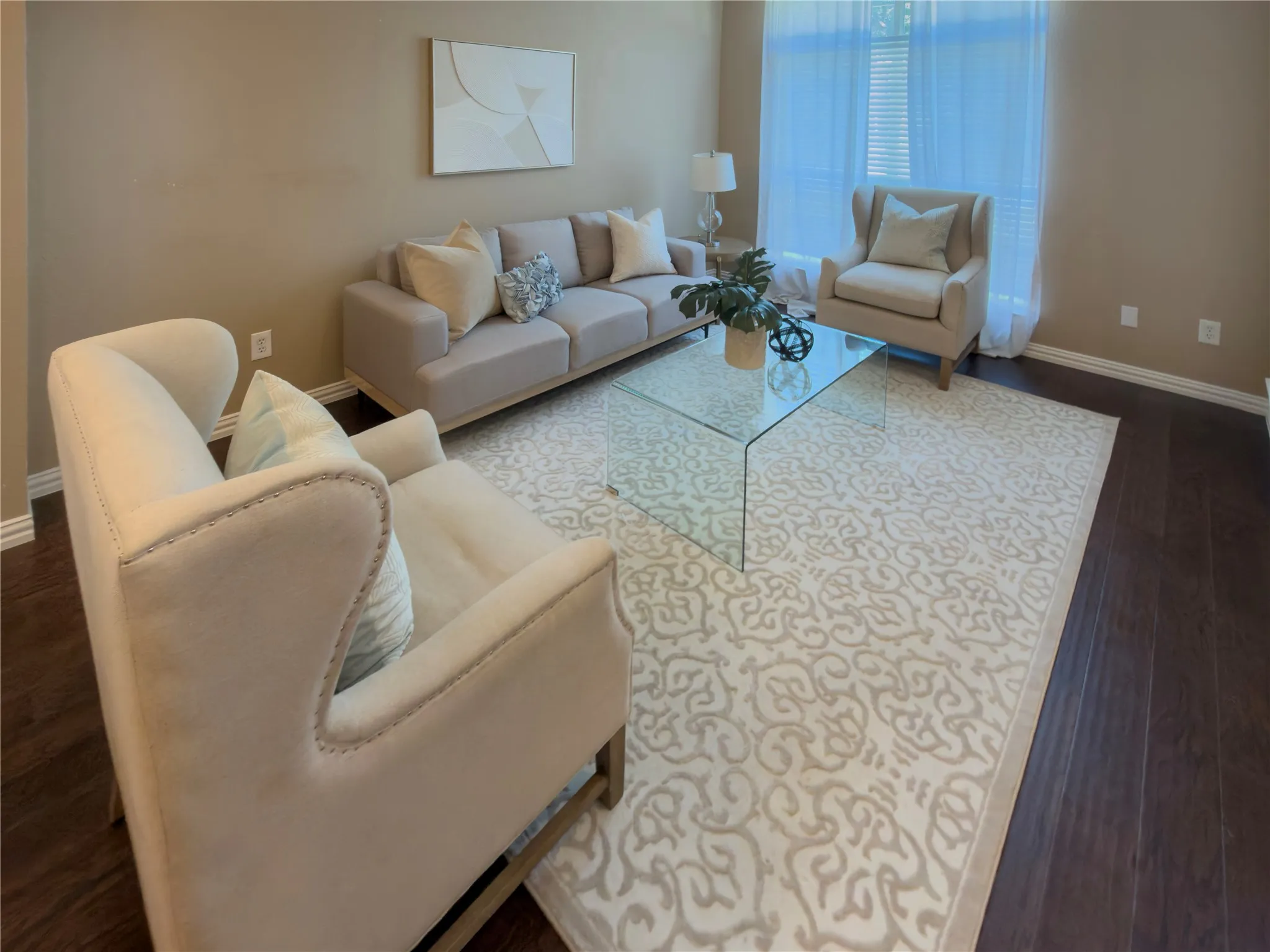 Dark wood-finish flooring, light tan wall color, large window with sheer white drapery, and a clear glass coffee table