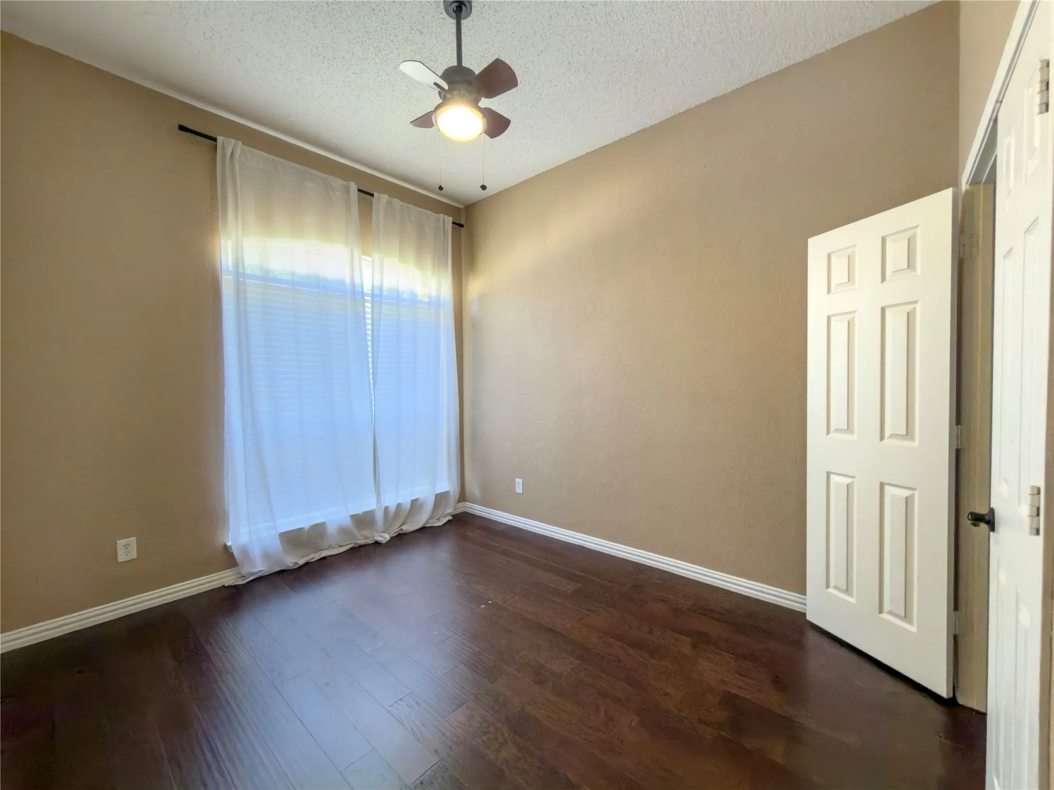 Rich wood-finish flooring, a large window with blinds, and a ceiling fan with integrated lighting