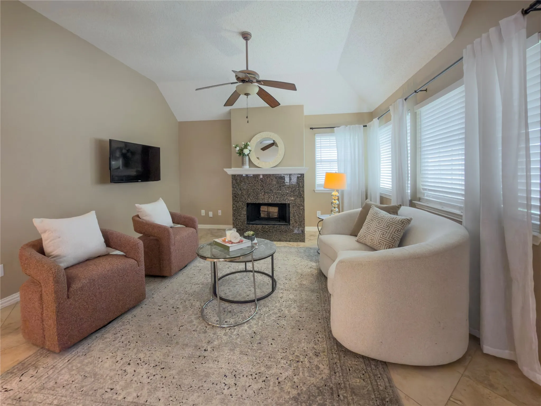 Vaulted ceiling with a ceiling fan, fireplace with a stone surround and mantle, and multiple windows with blinds and sheer curtains