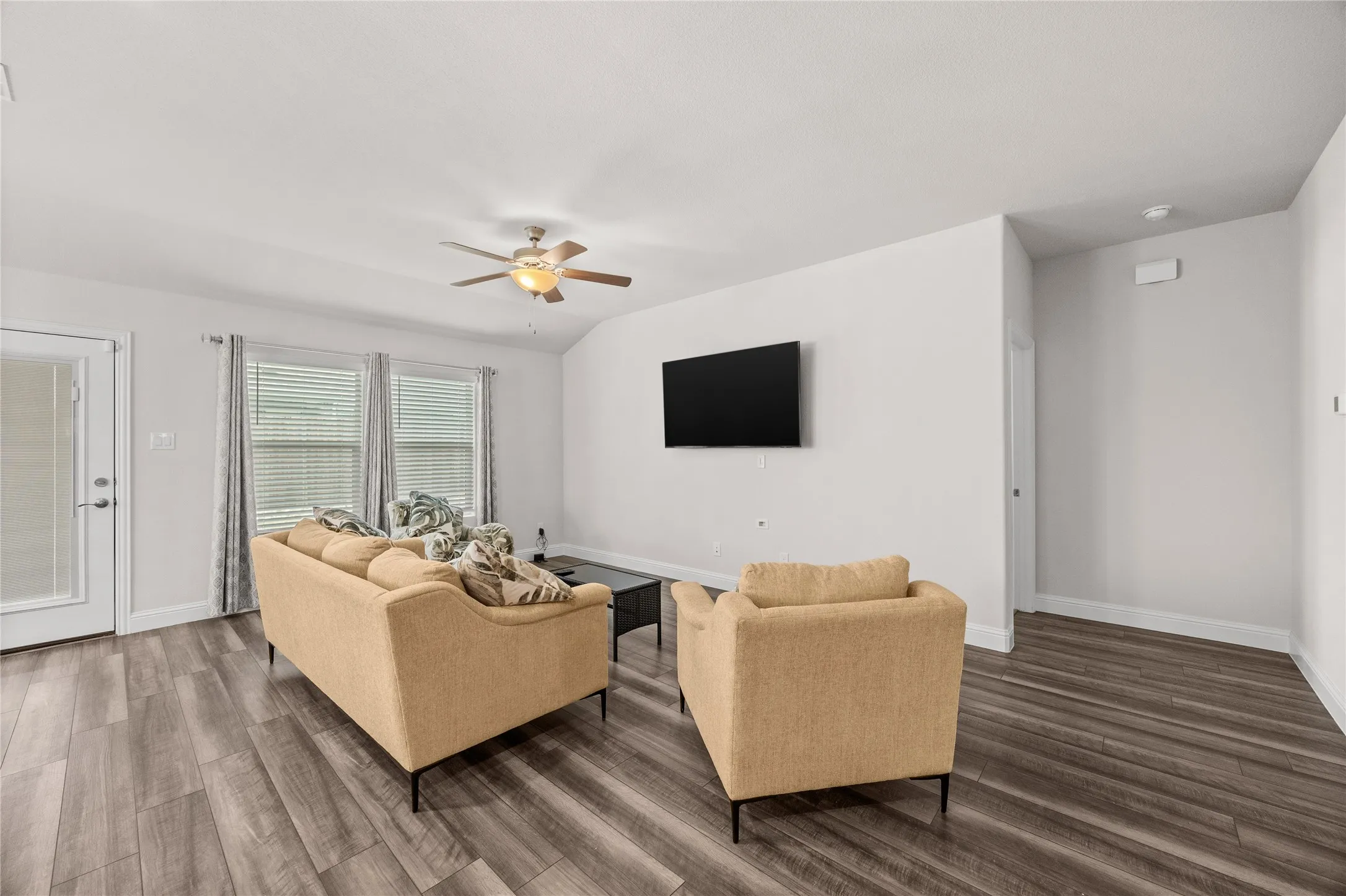 Wood-finish flooring throughout, vaulted ceiling with a mounted ceiling fan, multiple windows with blinds, and a white paneled door with a privacy glass insert