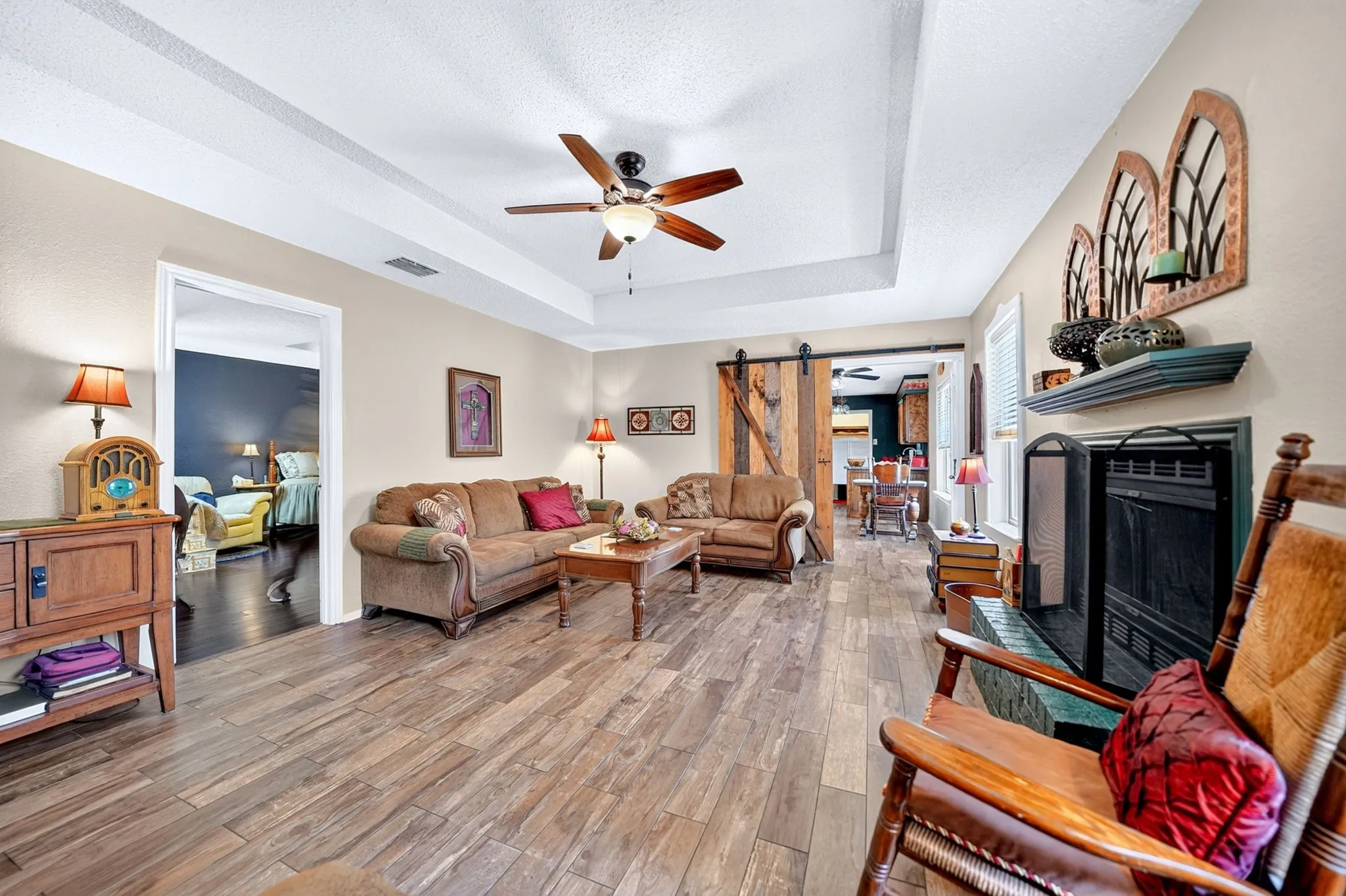 Tray ceiling with a ceiling fan, wood-finish flooring, fireplace with a mantel, and a sliding barn door