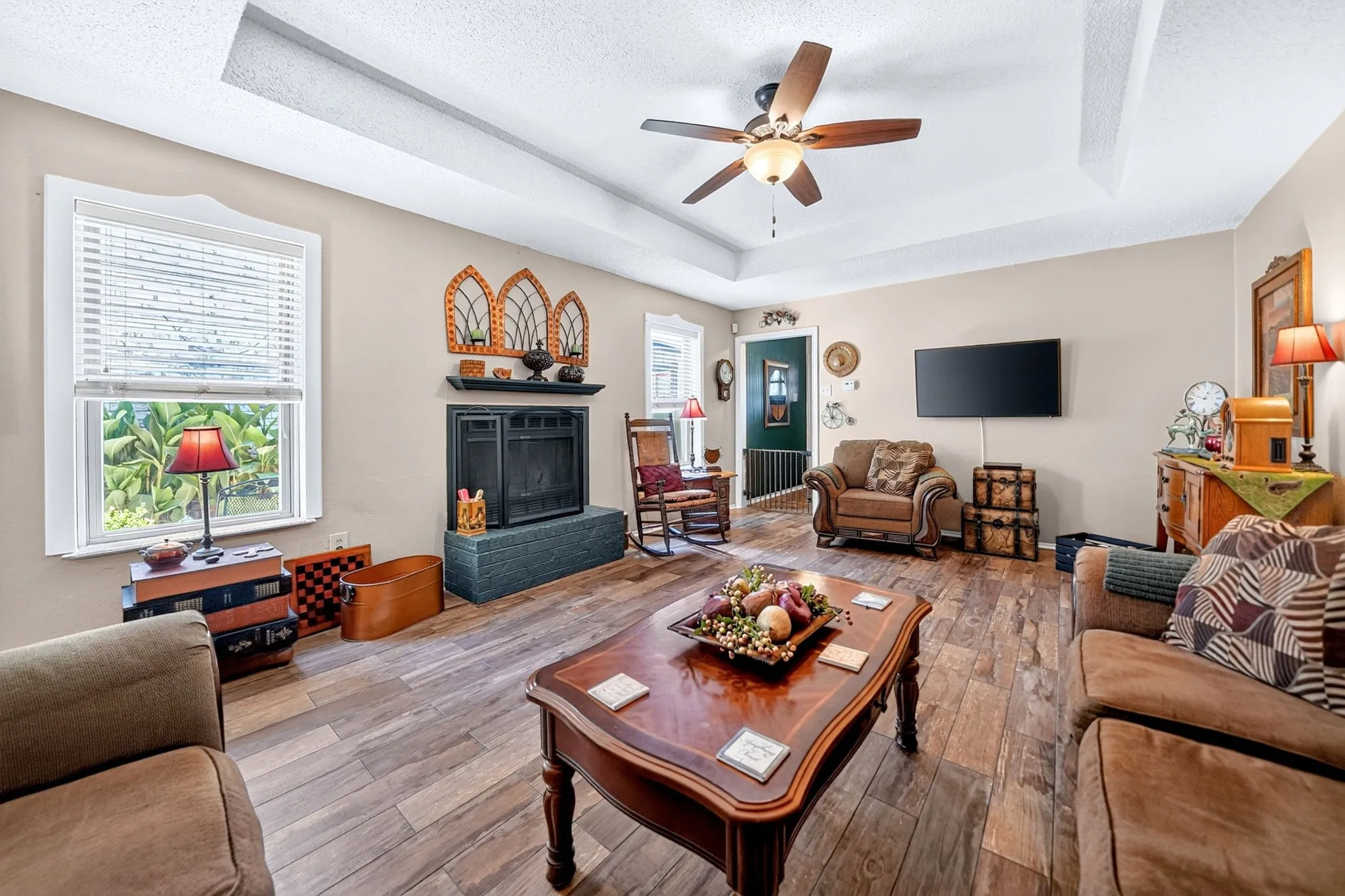 Tray ceiling with a ceiling fan, wood-finish flooring, a built-in fireplace with a dark mantel, and window with white blinds
