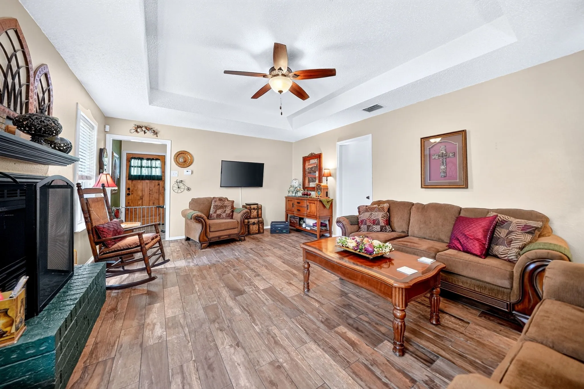 Tray ceiling, wood-finish flooring, ceiling fan, fireplace with brick surround, and paneled wood door