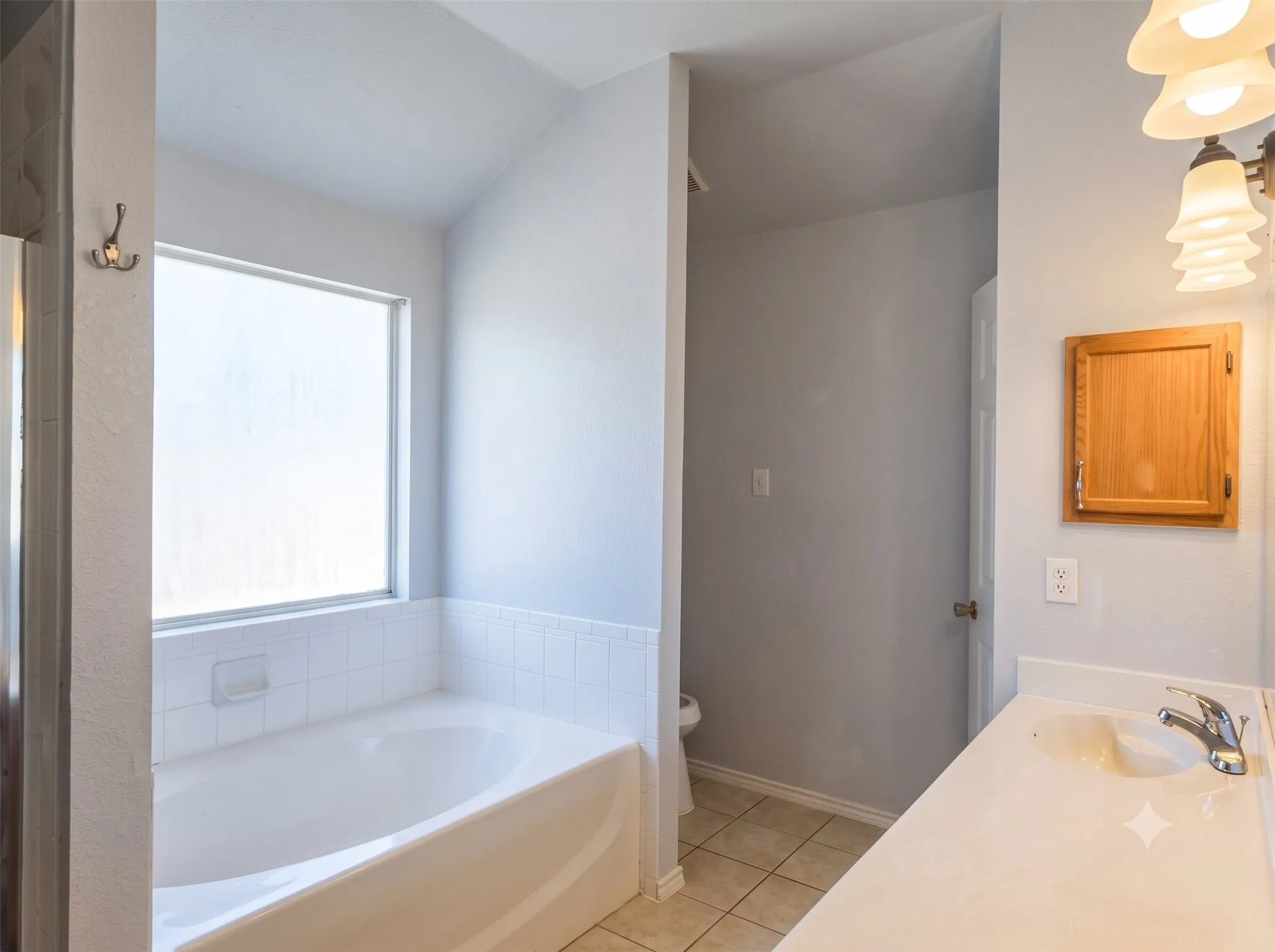 Soaking tub with tiled surround, large frosted window, vanity with integrated sink, and overhead lighting fixture