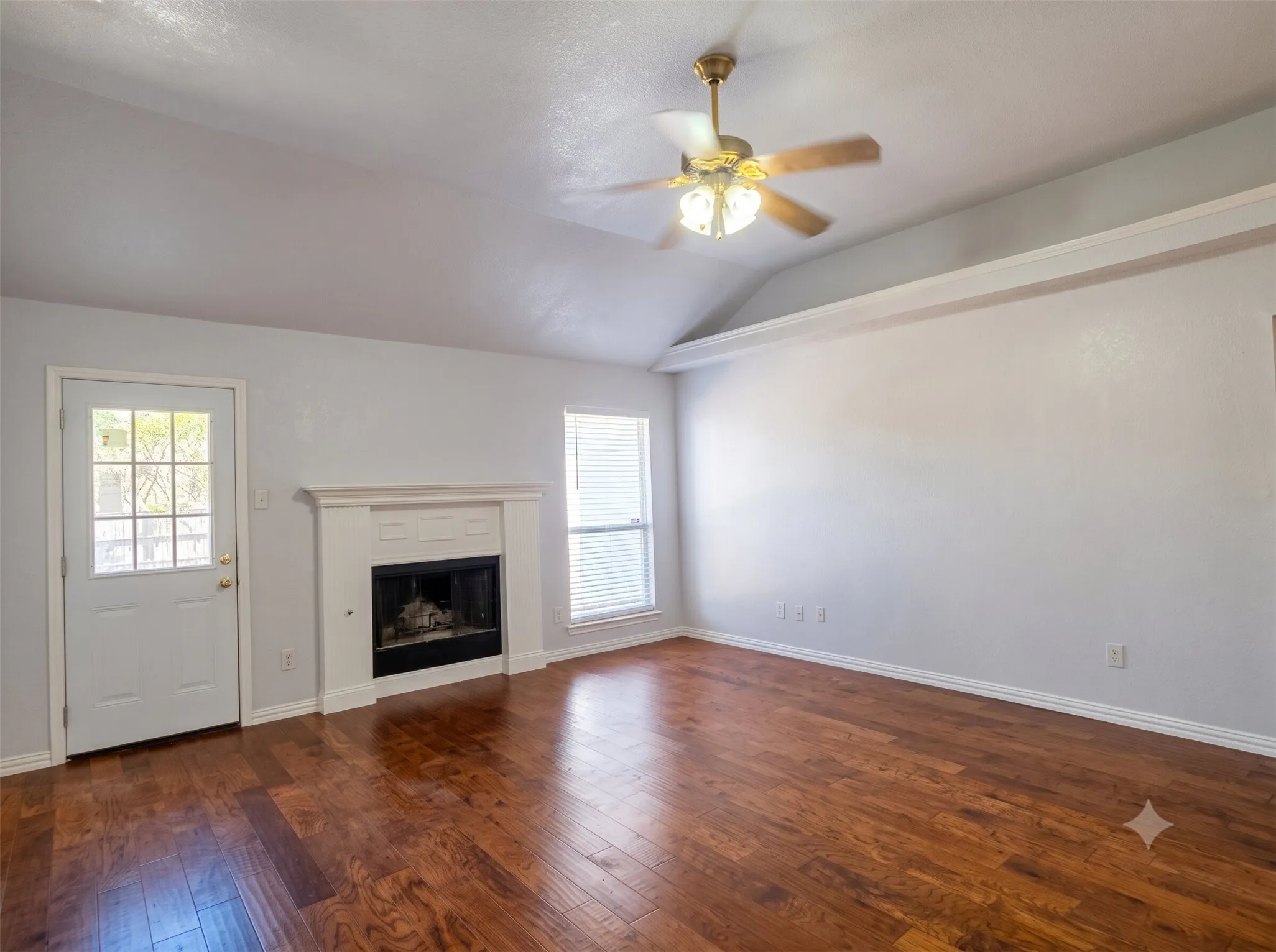 Wood-finish flooring, vaulted ceiling, fireplace with white mantel, ceiling fan with light fixture, and glass-paneled door
