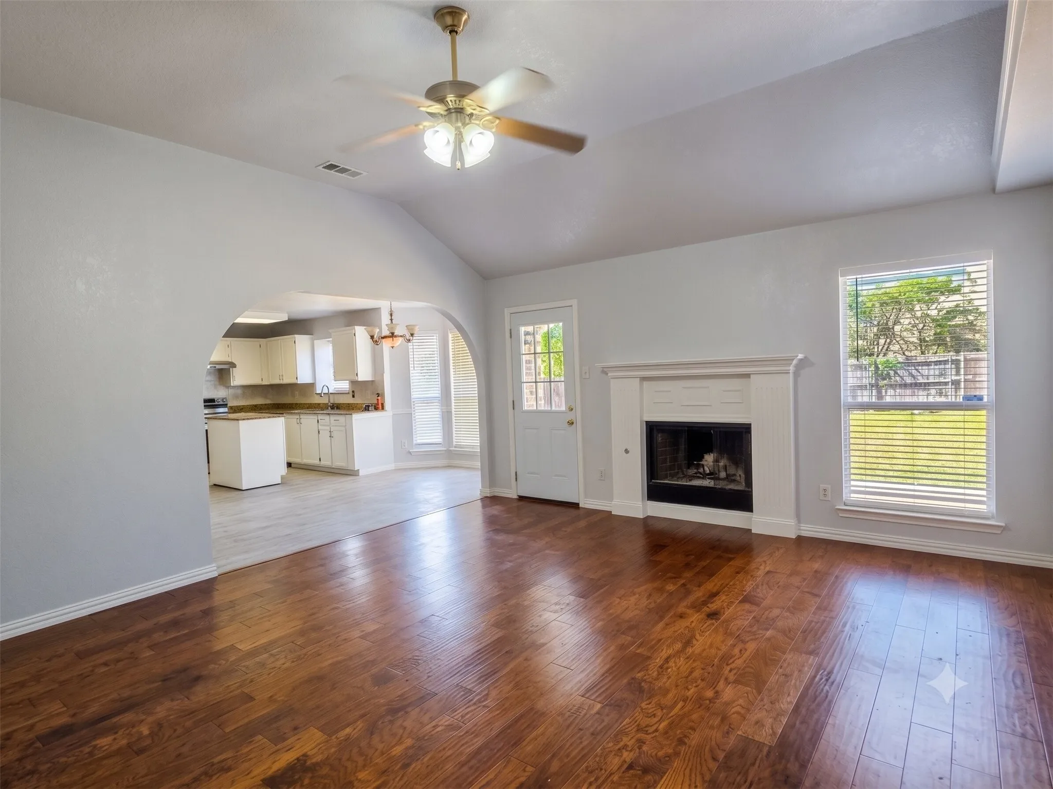 Vaulted ceiling with a ceiling fan, wood-finish flooring, white fireplace with a mantel, window with blinds, arched doorway