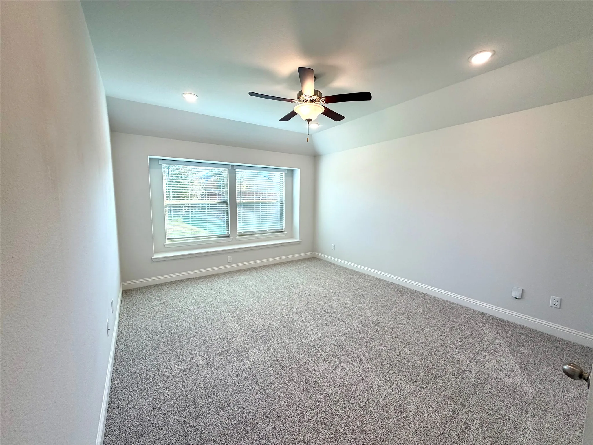 This spacious primary bedroom features a warm, neutral color palette throughout, complemented by plush, high-quality carpeting and modern recessed lighting. The room is equipped with a ceiling-mounted fan for climate comfort and features expansive windows outfitted with custom blinds that allow for an abundance of natural light. The well-proportioned layout provides generous wall space, offering ideal versatility for furniture placement and interior decor arrangements.