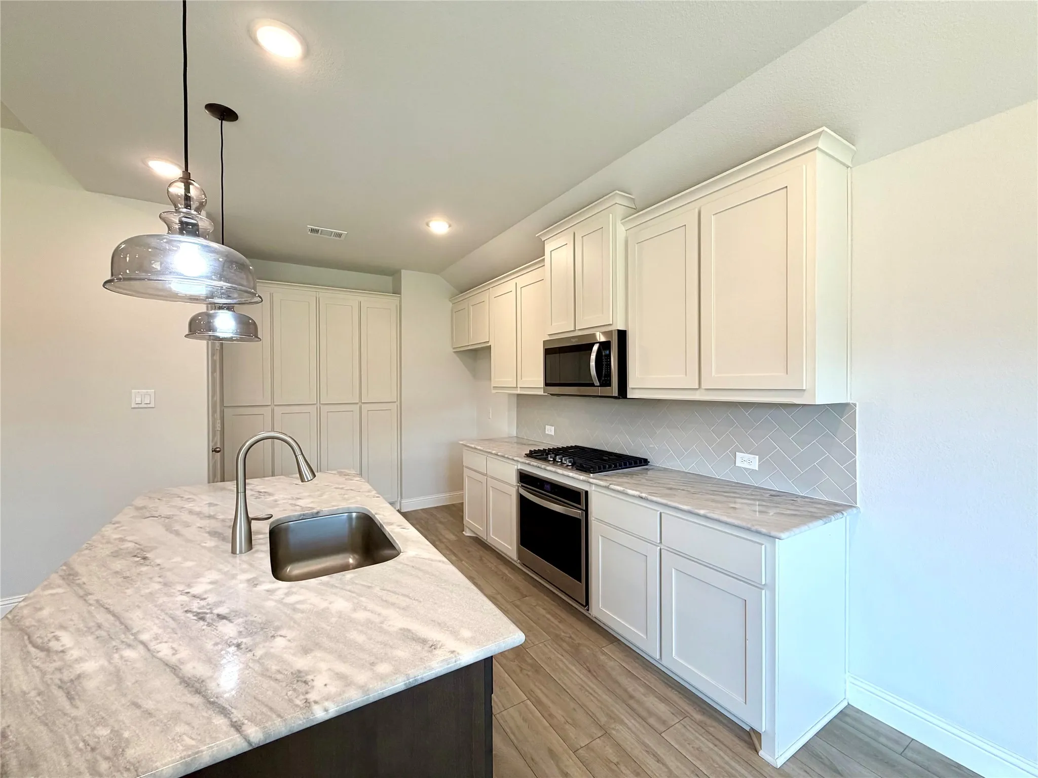 This modern, gourmet kitchen features elegant white shaker cabinetry paired with premium stainless steel appliances and a stylish herringbone tile backsplash. The heart of the room is a spacious kitchen island that includes a convenient built-in sink, all complemented by durable, high-end luxury vinyl plank (LVP) flooring throughout the space.