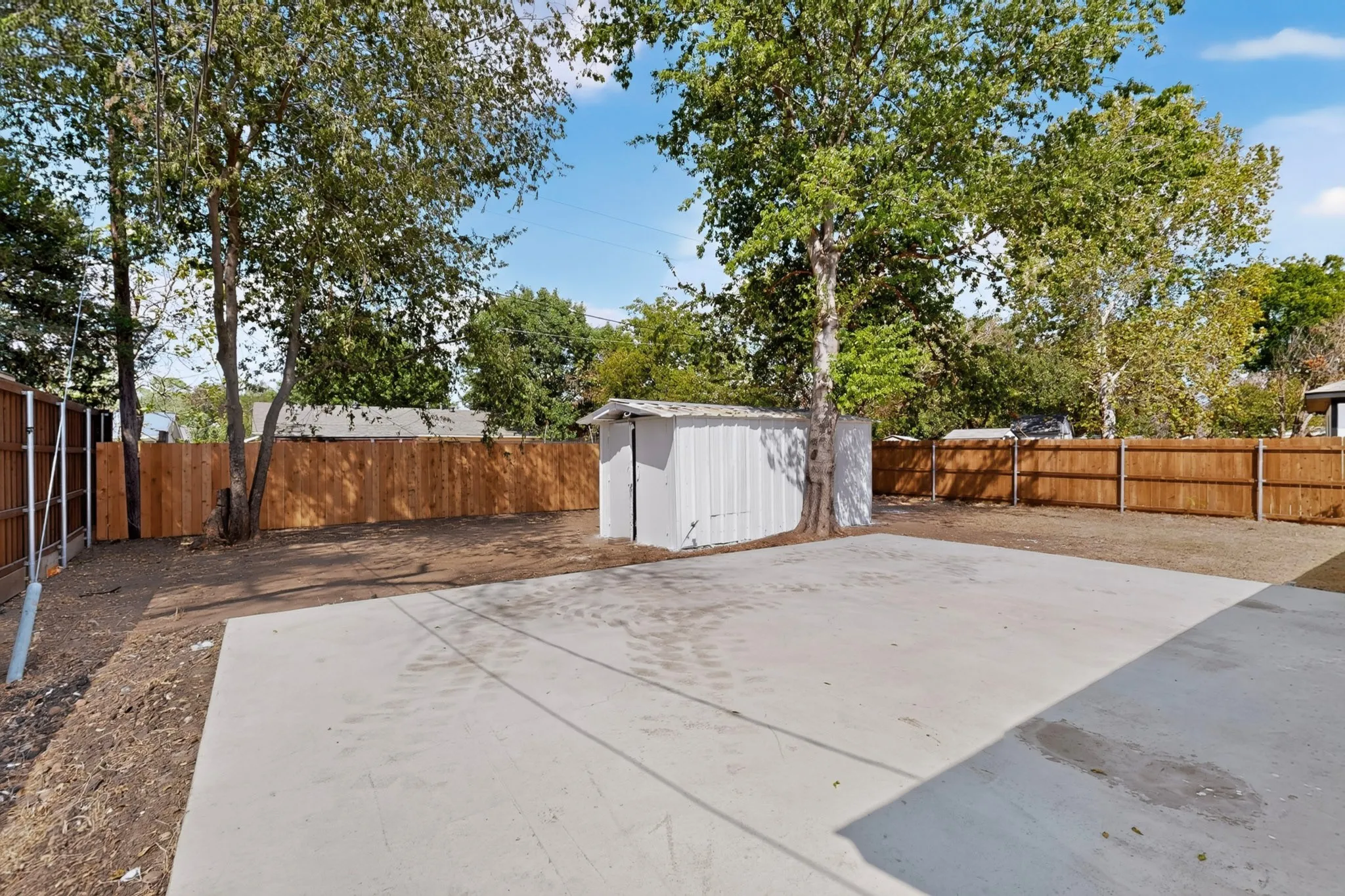 Expansive concrete patio with a storage shed, mature trees, and wood privacy fencing
